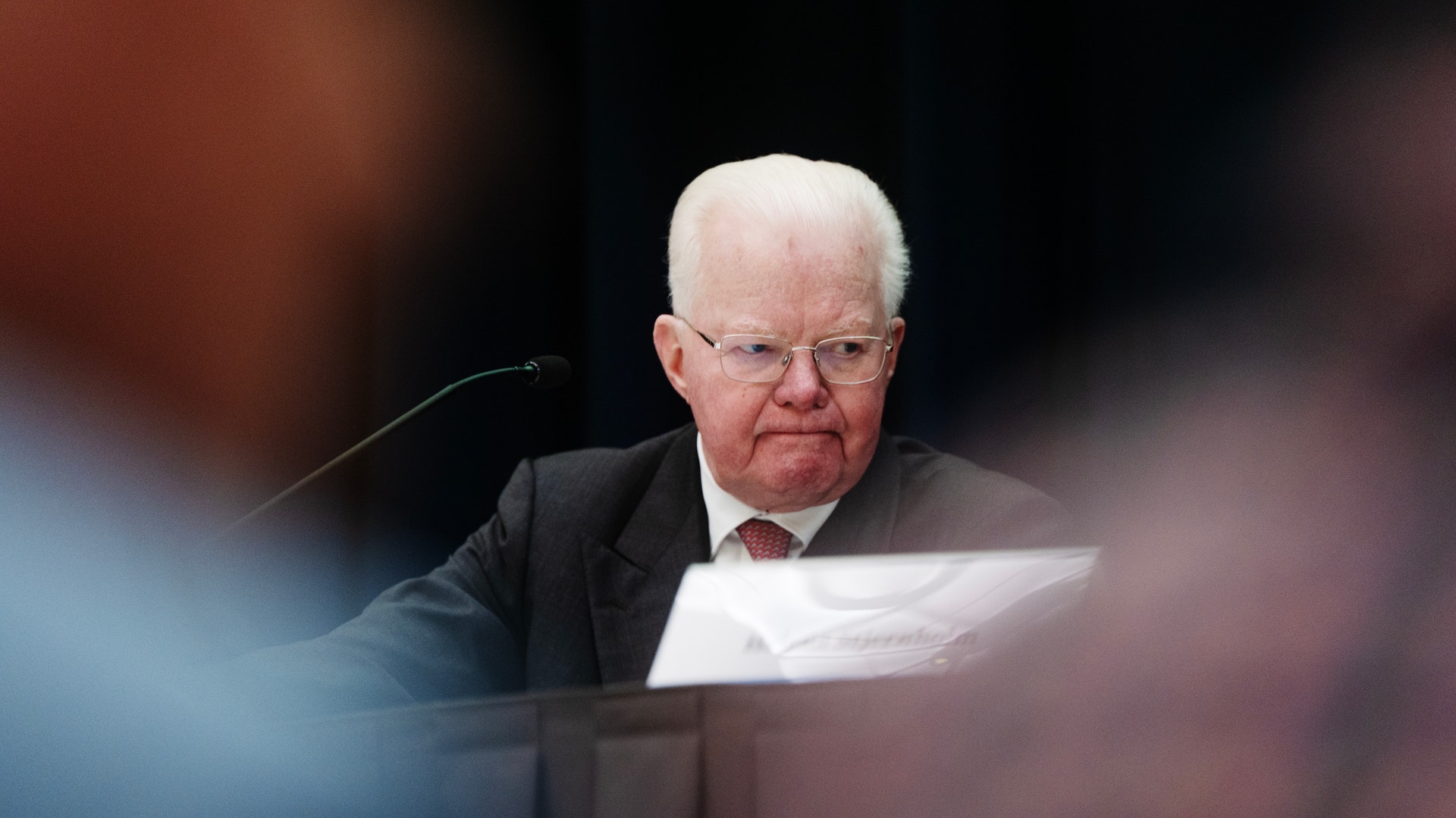 En äldre man i kostym och glasögon talar vid ett podium med mikrofon framför en suddig publik.