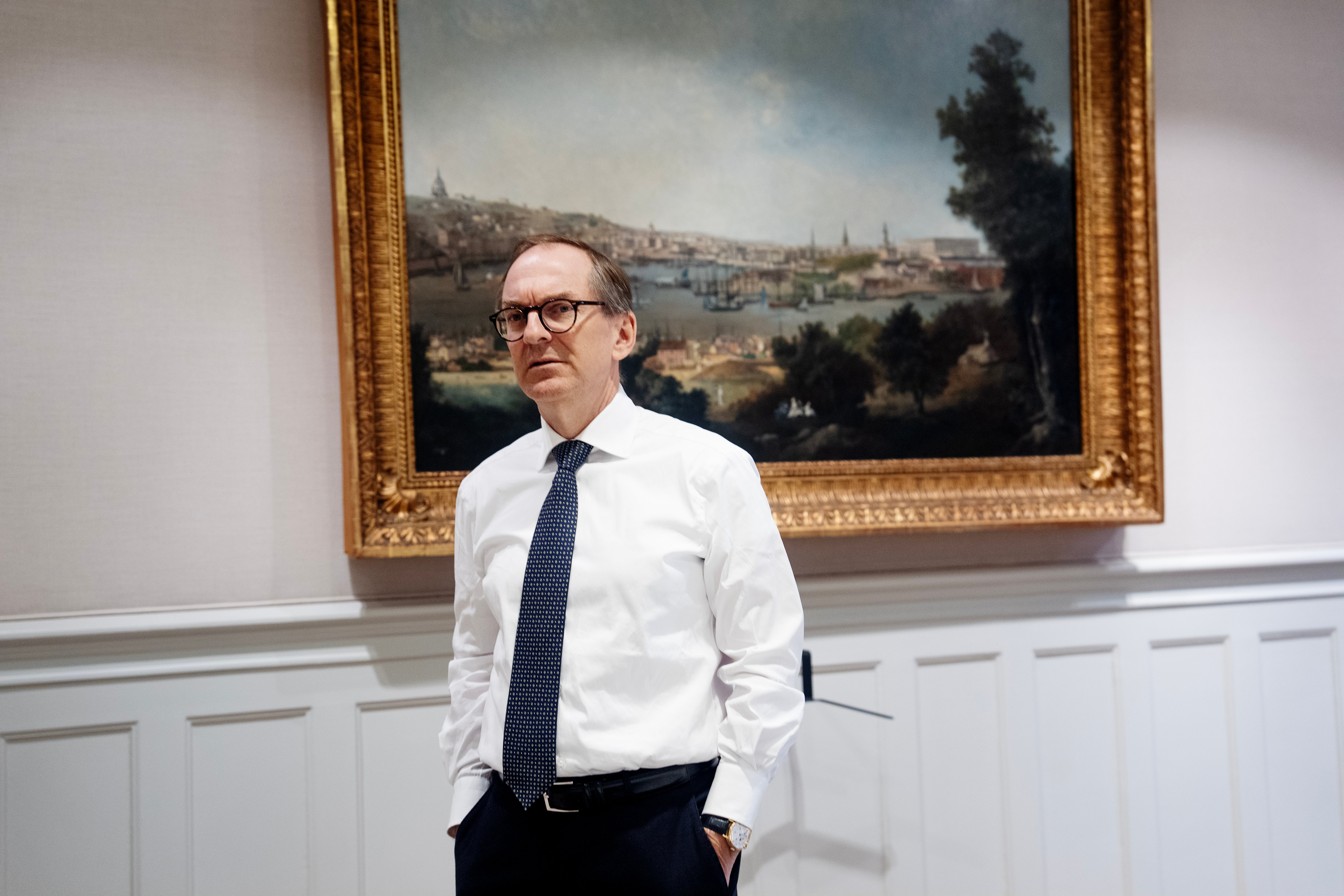 Porträttfoto av en man i vit skjorta och mörk slips som står framför en målad landskapsbild i en elegant inredning.