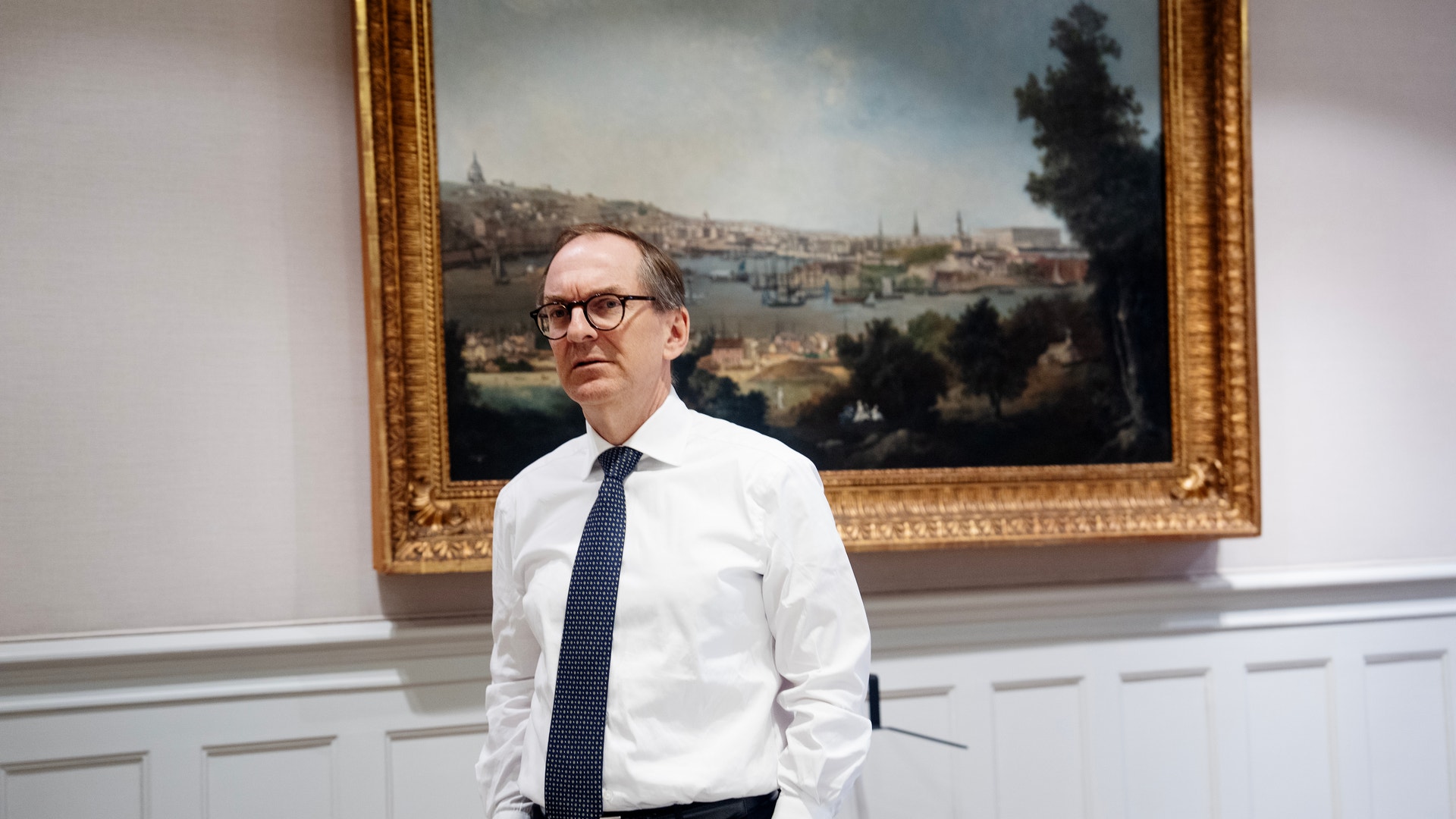 Porträttfoto av en man i vit skjorta och mörk slips som står framför en målad landskapsbild i en elegant inredning.