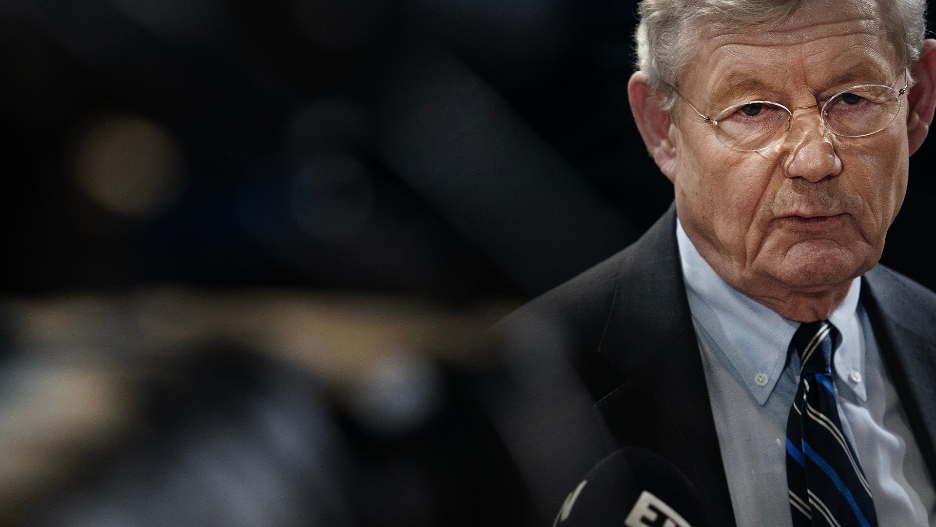 Äldre man med glasögon i mörk kostym talar vid ett officiellt tillfälle.