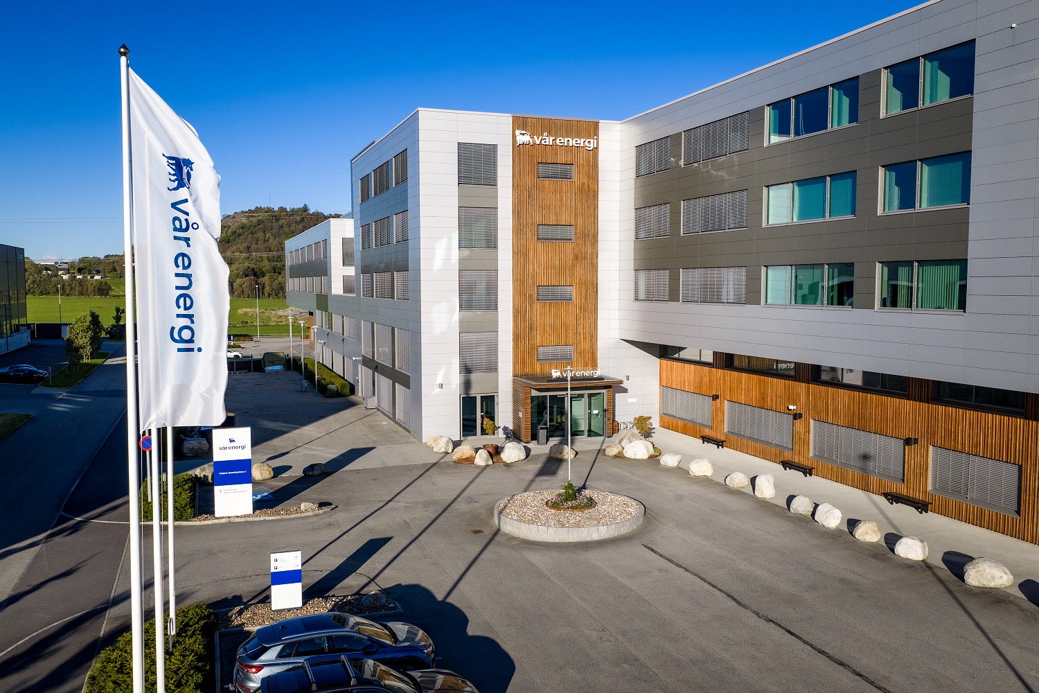 Modernt kontorsbyggnad med vita och träfärgade fasader, flagga i förgrunden och öppen plats framför entrén.