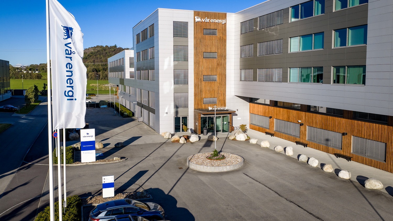 Modernt kontorsbyggnad med vita och träfärgade fasader, flagga i förgrunden och öppen plats framför entrén.