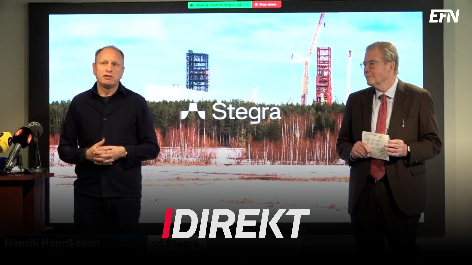 Två män i en TV-studio diskuterar ett byggprojekt. I bakgrunden syns en konstruktionsplats med kranar och byggnader.