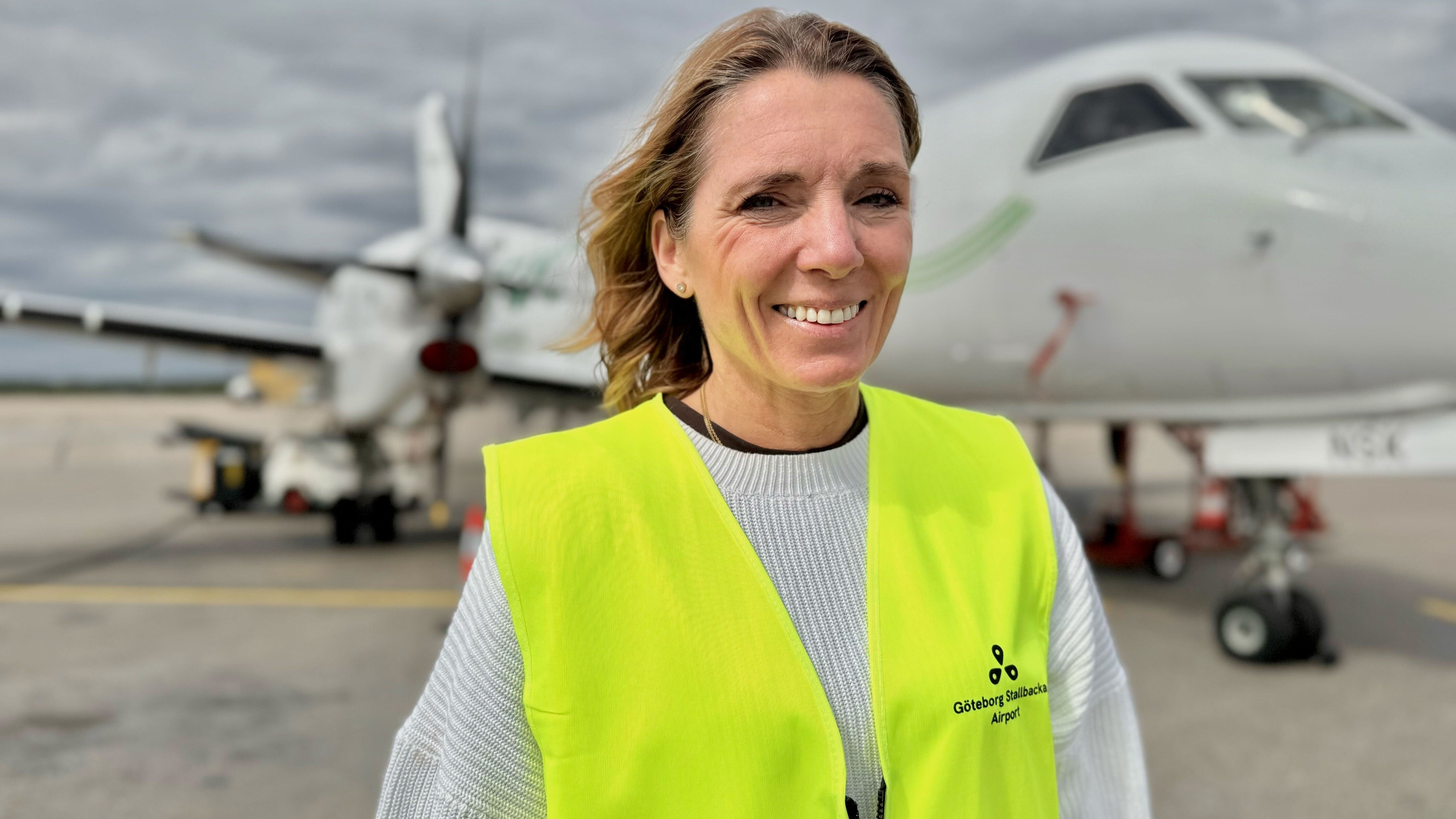 Porträttfoto av en leende kvinna i gul säkerhetsvest framför ett passagerarflygplan på en flygplats.