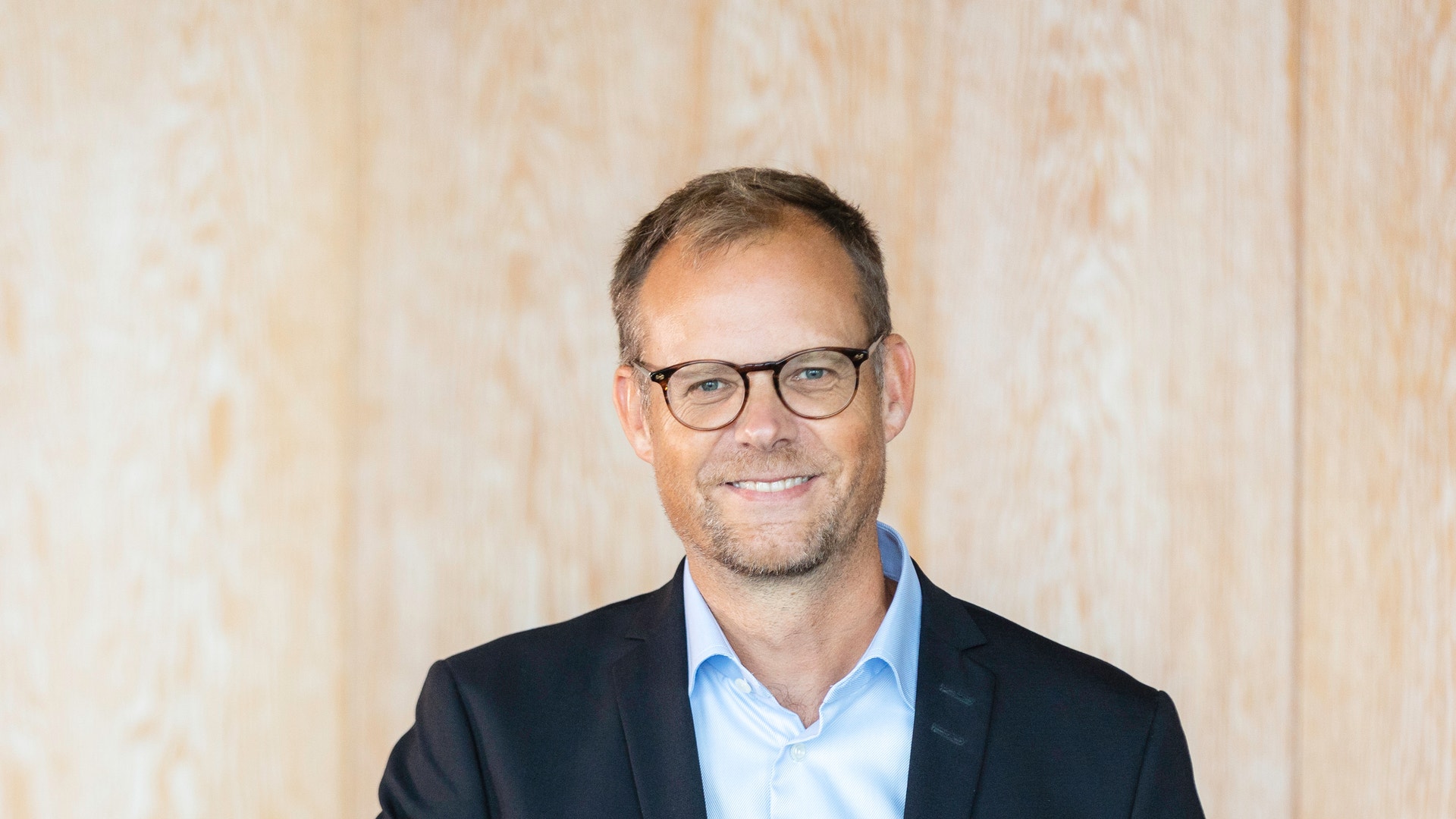 Porträttfoto av en man i svart blazer och ljusblå skjorta sittande vid ett bord med en kaffekopp