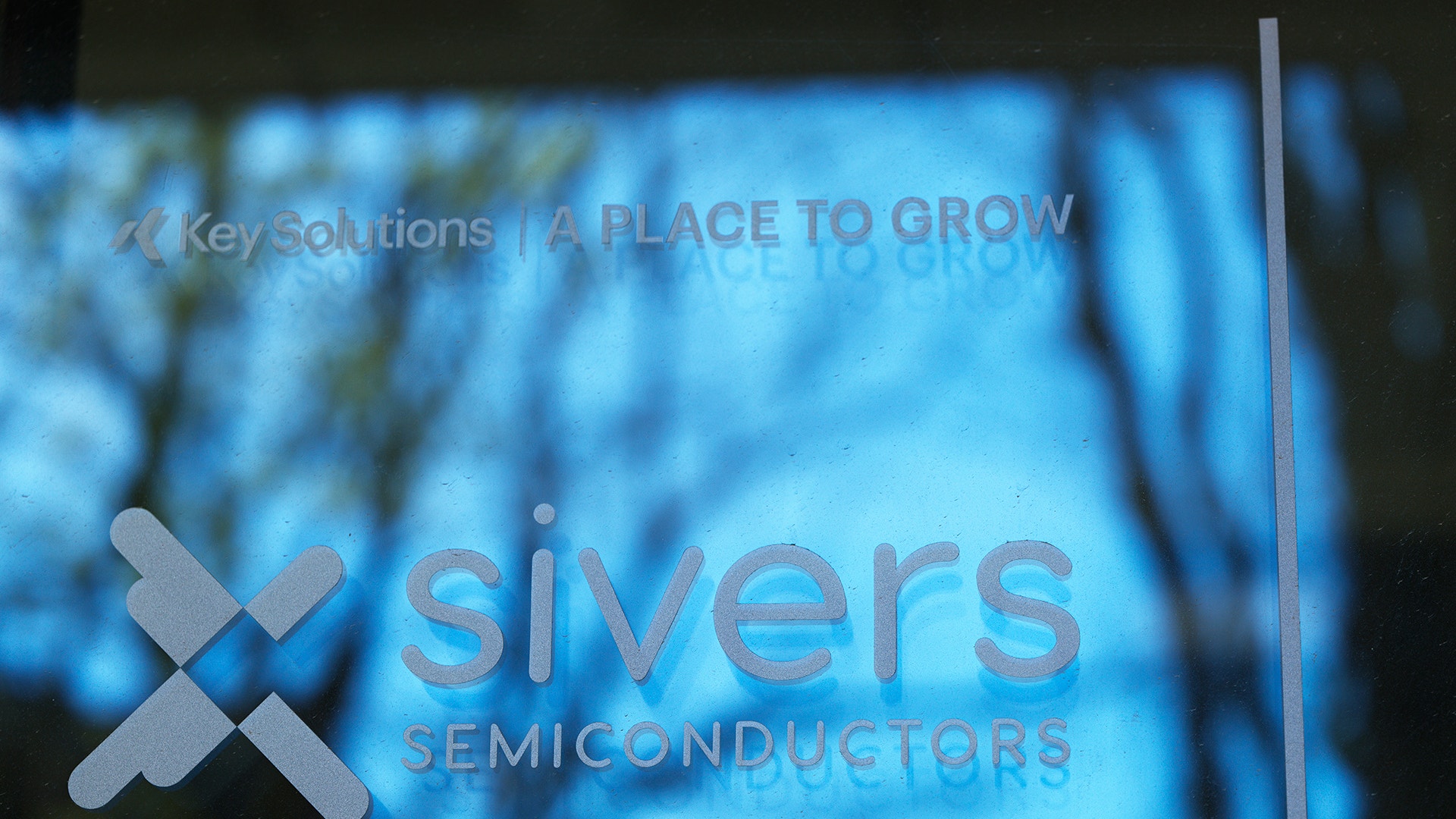 Sivers Semiconductors logotyp och text på glasyta med blå bakgrundsbelysning