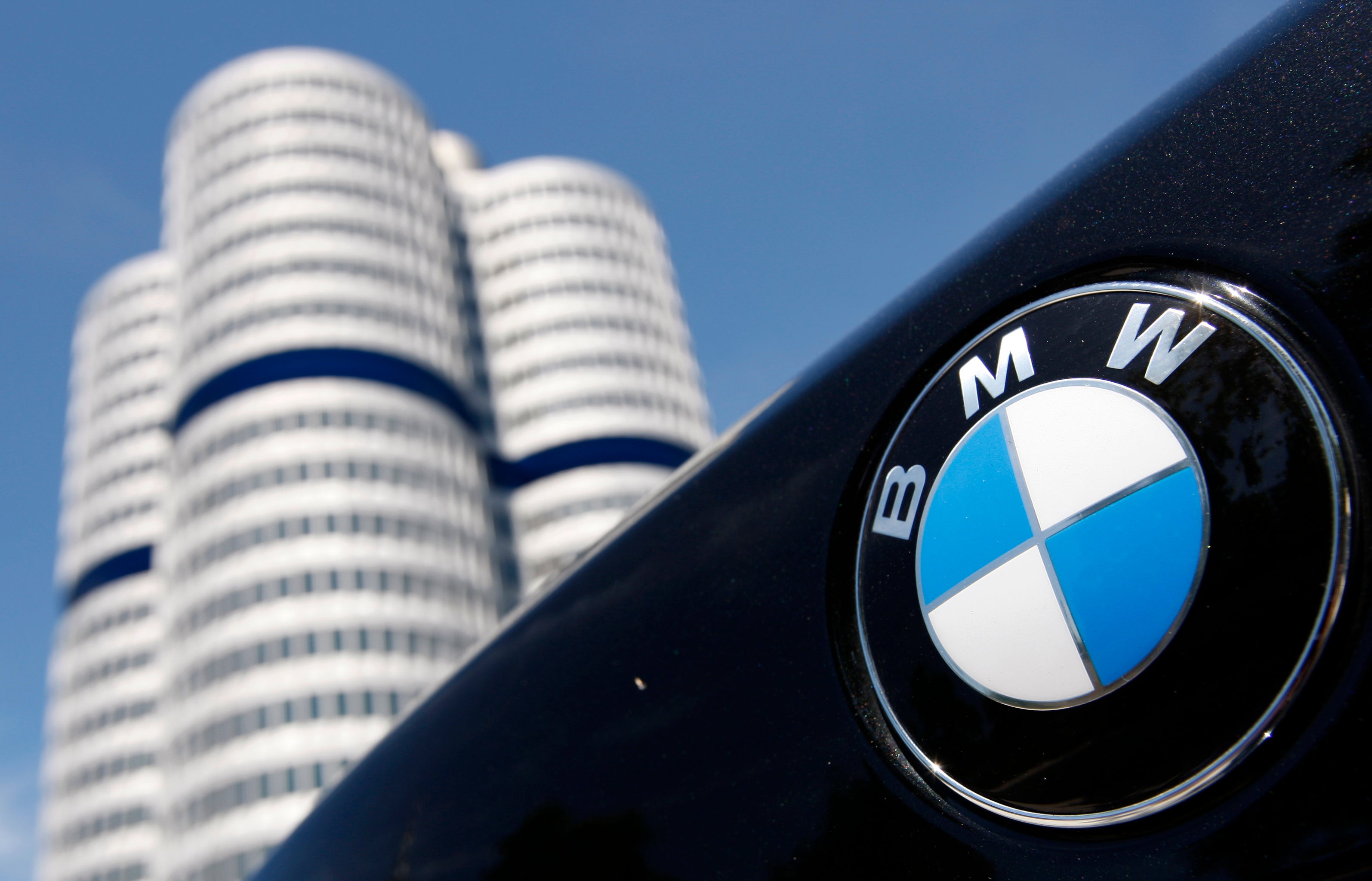 BMW-logotypen på en bil med BMW:s huvudkontor i form av vita cylindriska torn i bakgrunden mot blå himmel.