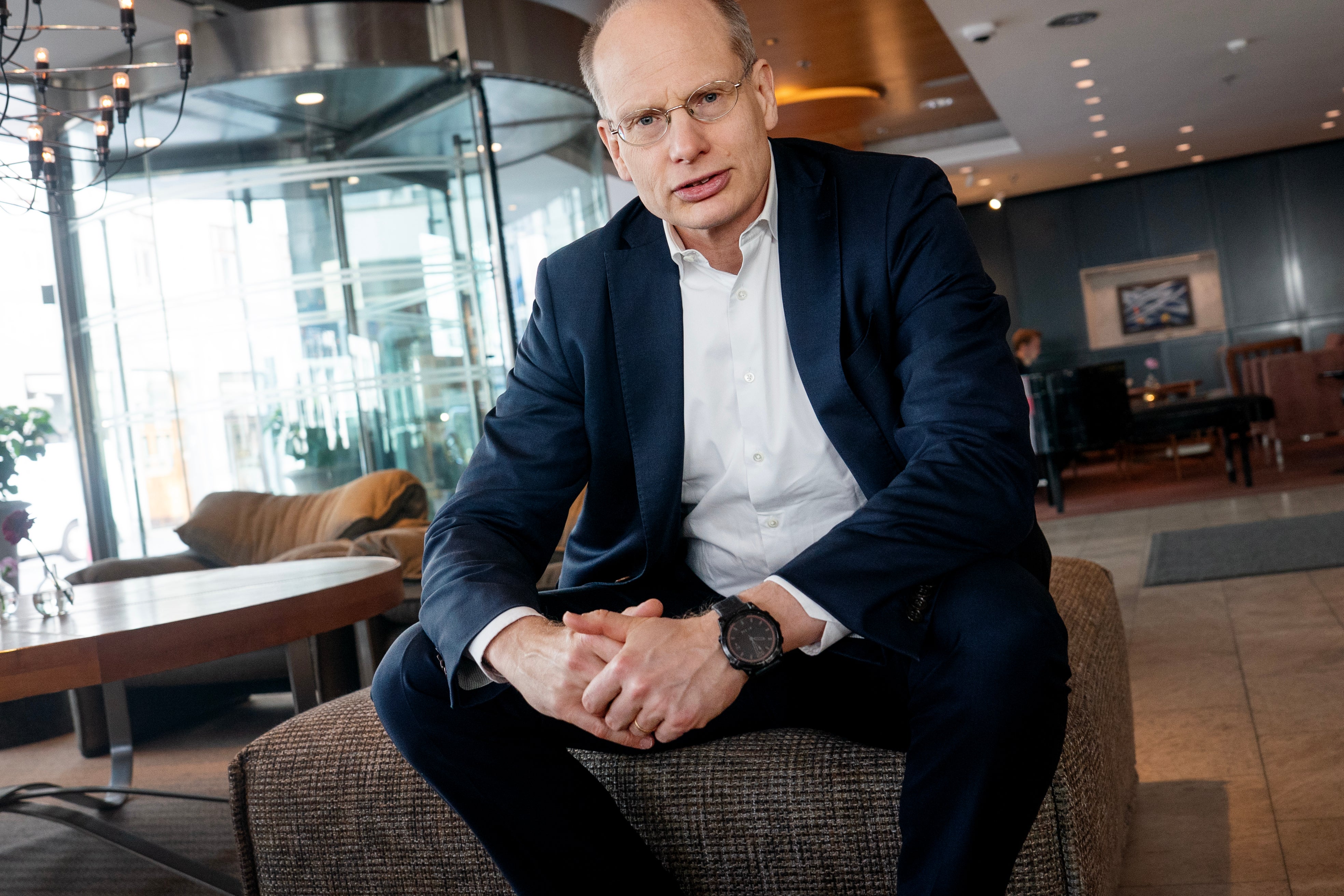 Man i mörkblå kostym och vit skjorta sitter på en stol i en modern kontorsmiljö med glasvägg och belysning i bakgrunden.