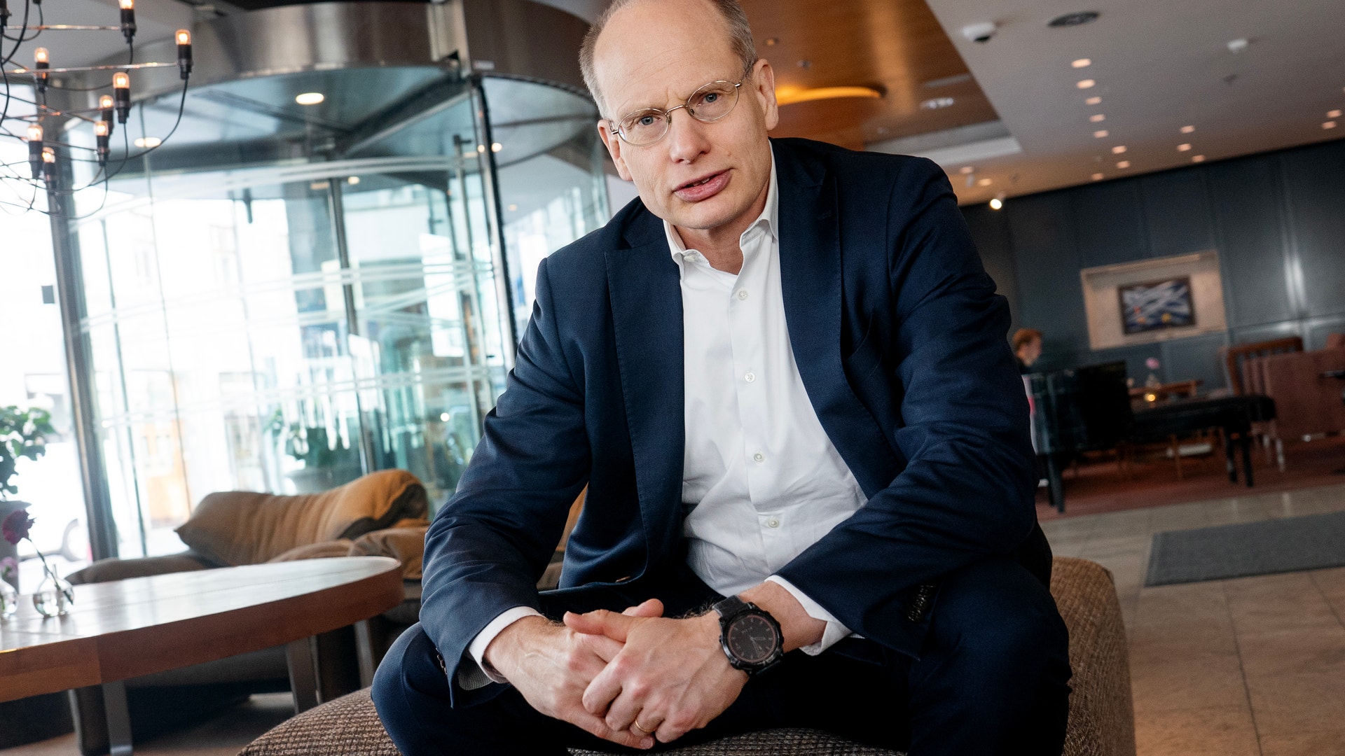Man i mörkblå kostym och vit skjorta sitter på en stol i en modern kontorsmiljö med glasvägg och belysning i bakgrunden.