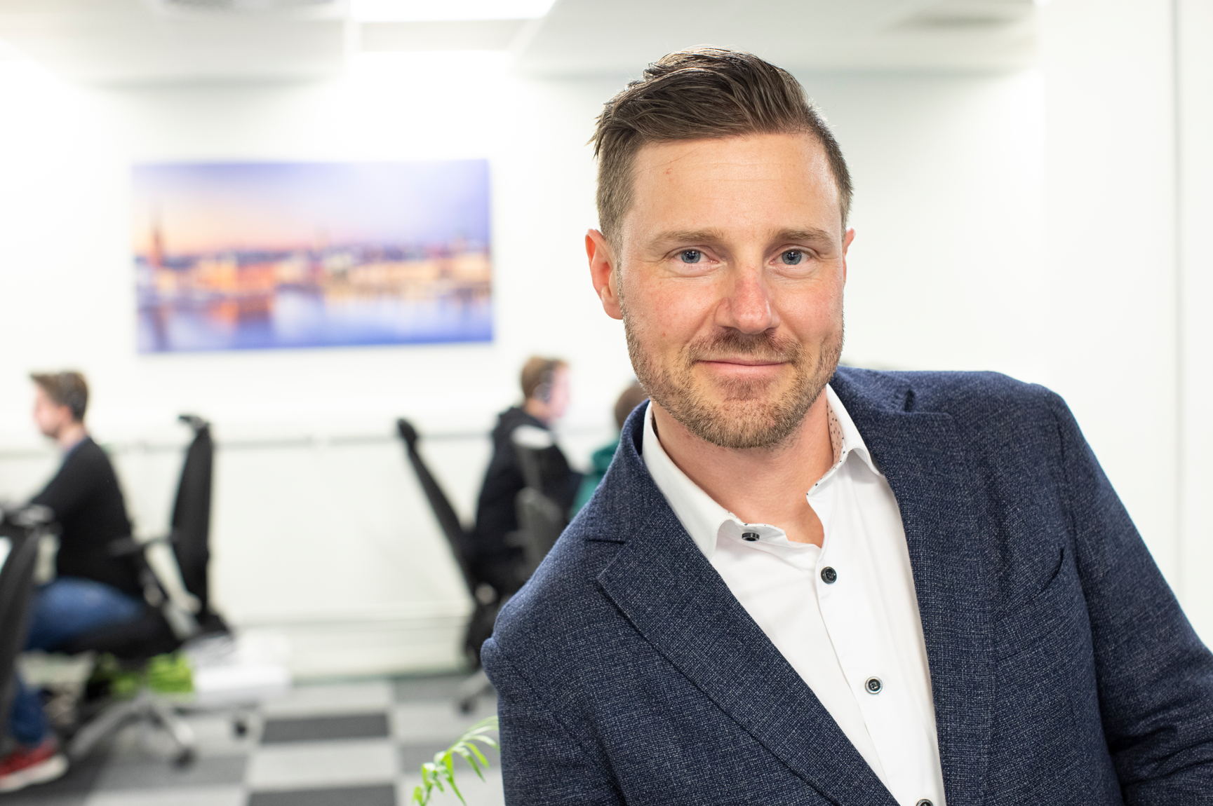 Man i kavaj ler mot kameran på ett kontor, med kollegor som arbetar i bakgrunden.