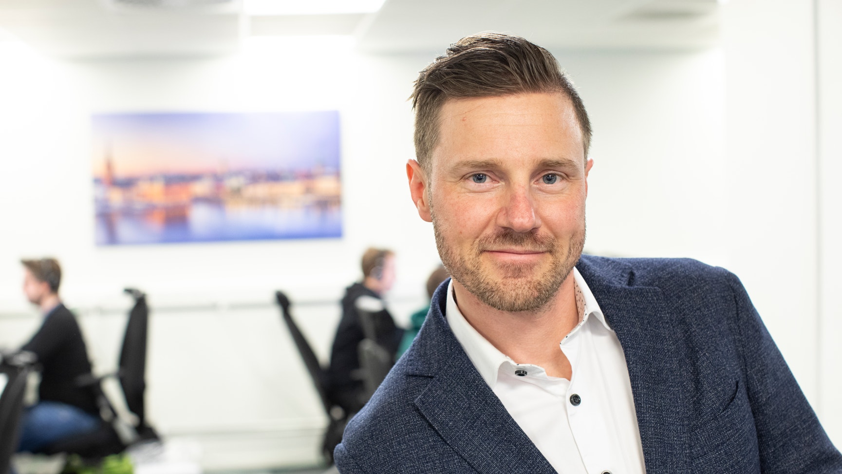 Man i kavaj ler mot kameran på ett kontor, med kollegor som arbetar i bakgrunden.