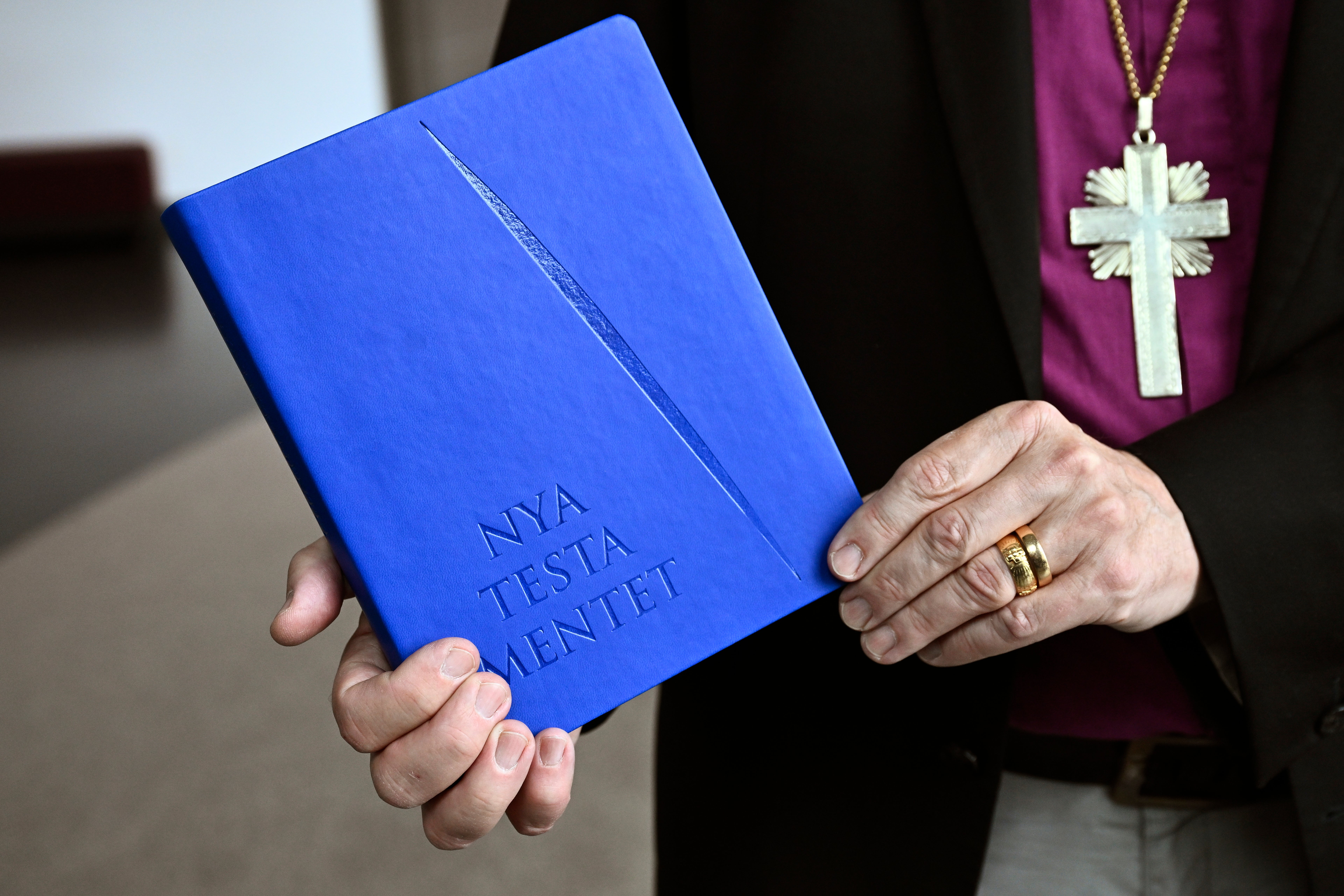 En person i prästkläder med kors håller i en blå bok med texten Nya Testamentet.