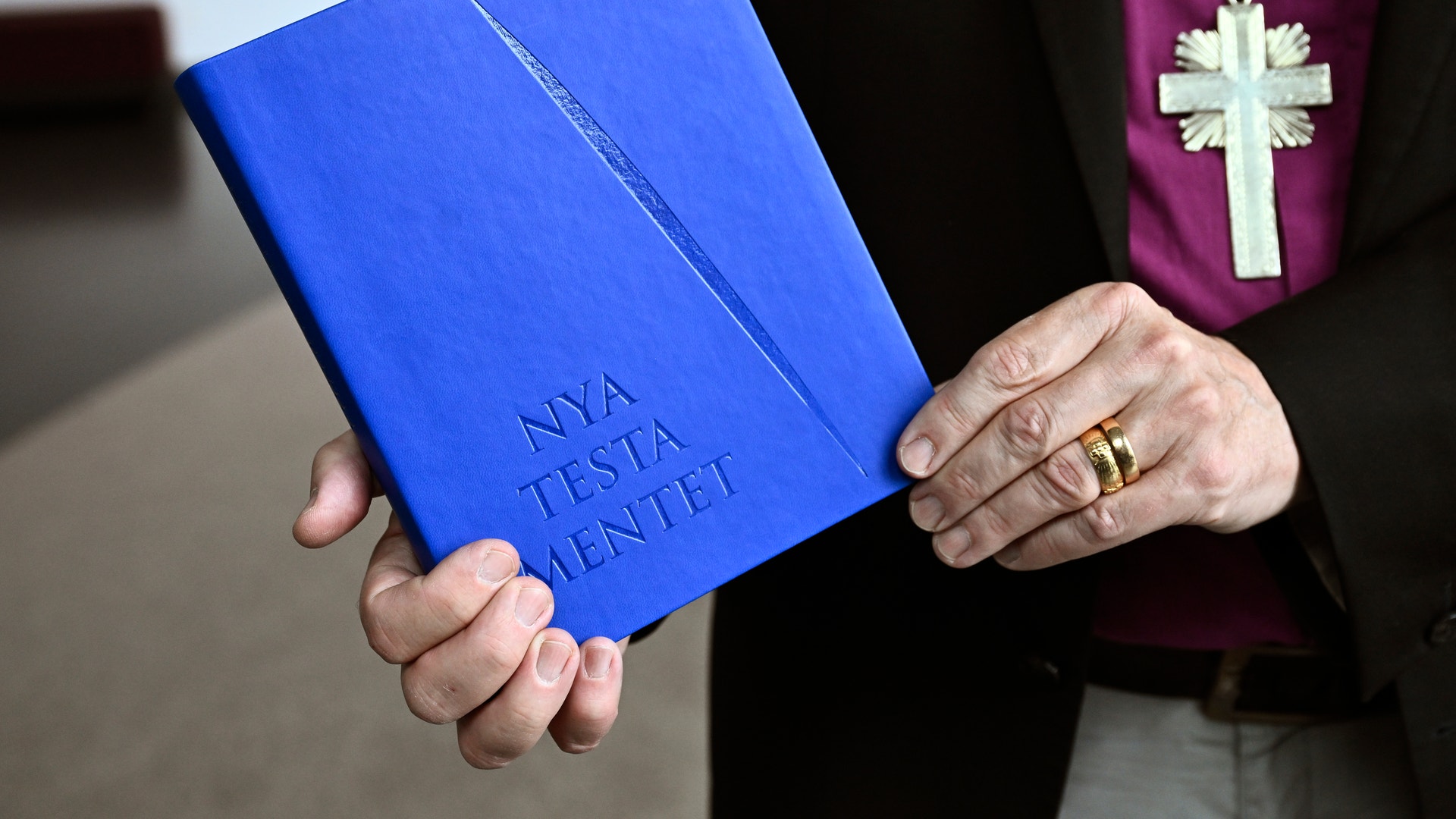 En person i prästkläder med kors håller i en blå bok med texten Nya Testamentet.