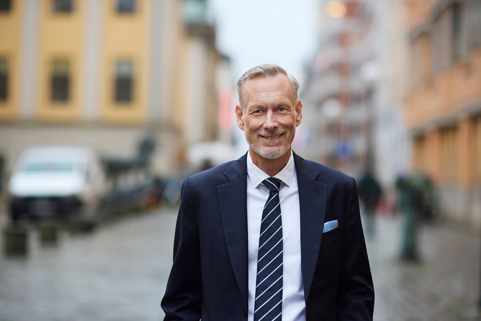Leende man i mörk kostym och randig slips som står utomhus på en gata i staden.