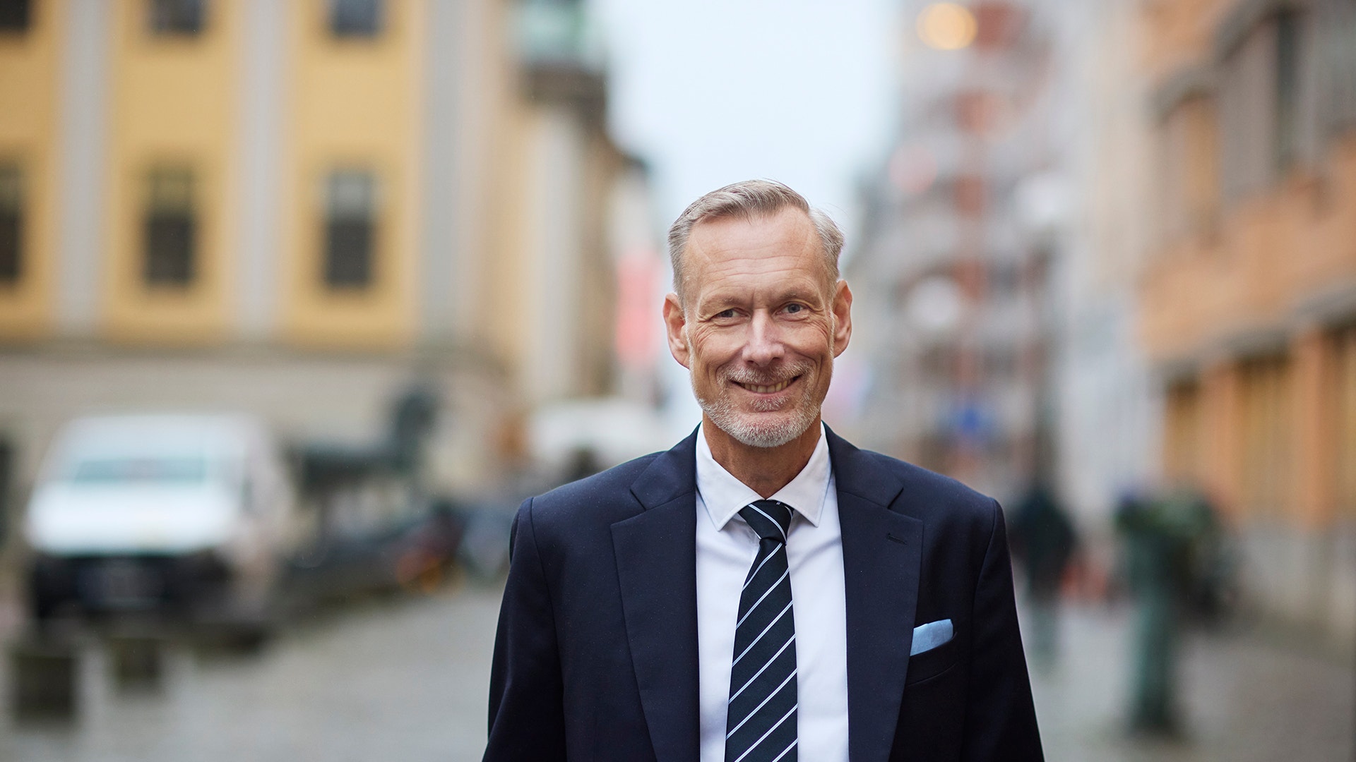 Leende man i mörk kostym och randig slips som står utomhus på en gata i staden.