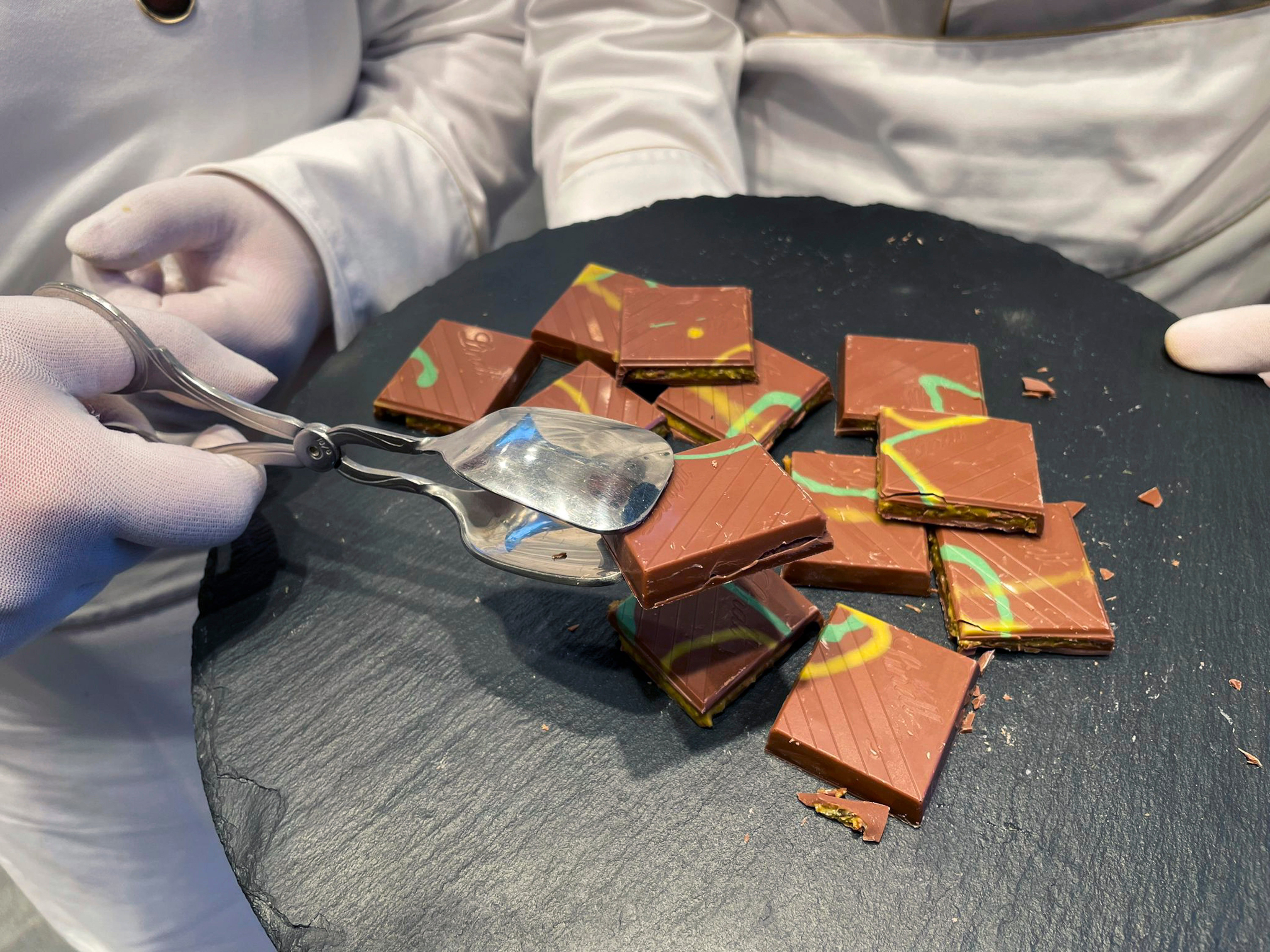 En person i vita handskar serverar bitar av chokladkaka med färgglada dekorationer från en rund skiffertallrik.