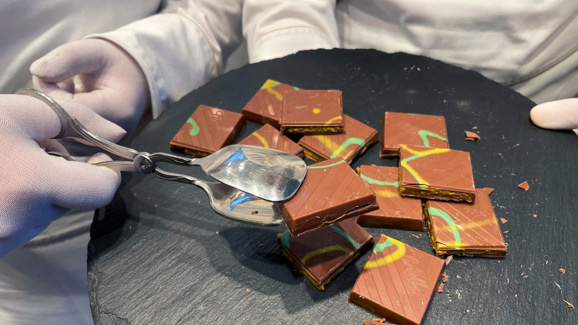 En person i vita handskar serverar bitar av chokladkaka med färgglada dekorationer från en rund skiffertallrik.