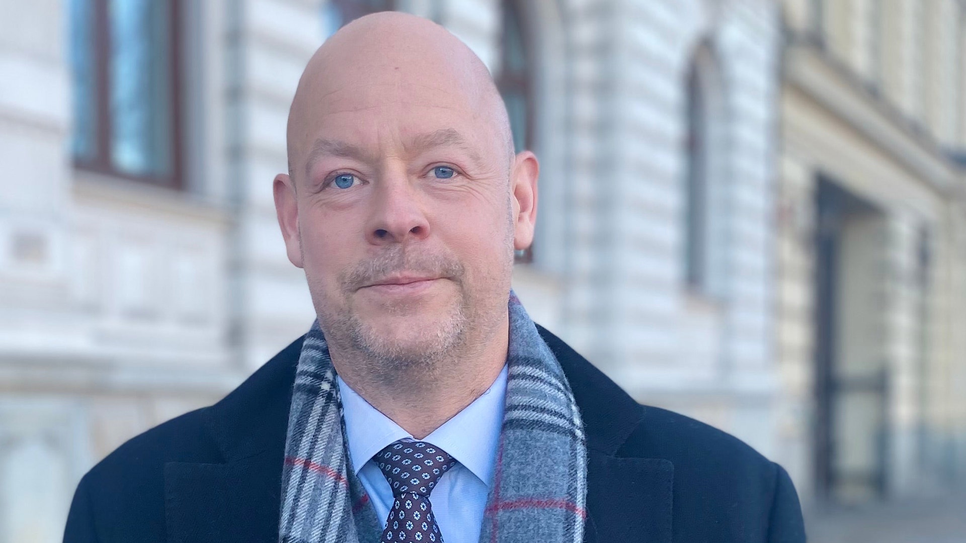 Porträtt av en man i mörkblå kavaj, ljusblå skjorta och slips mot en stadsbakgrund.