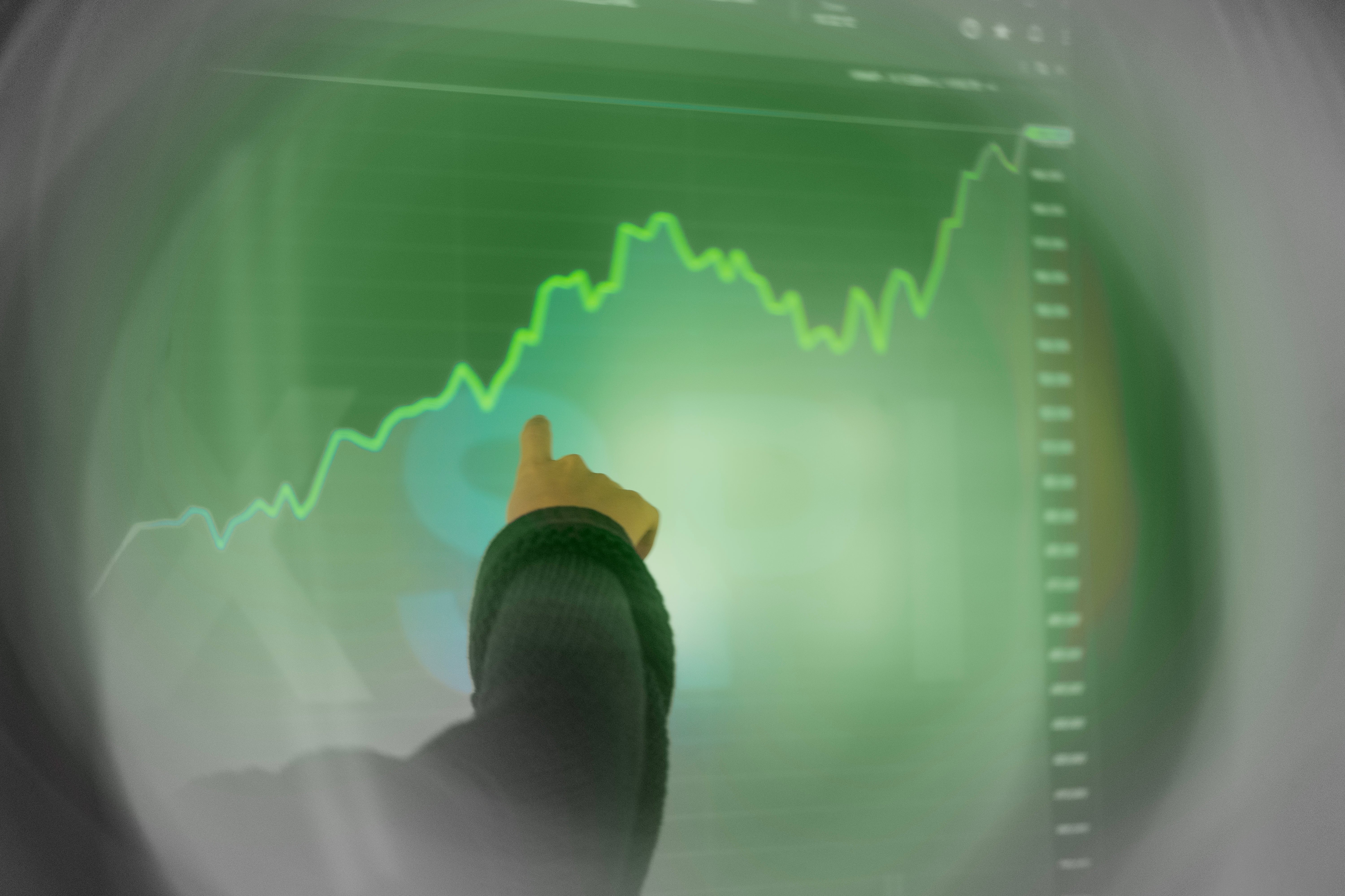 Person som tittar på en stigande finansiell graf med gröna trendlinjer på en skärm.