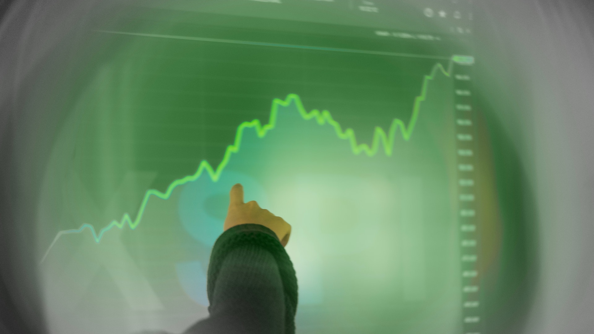 Person som tittar på en stigande finansiell graf med gröna trendlinjer på en skärm.
