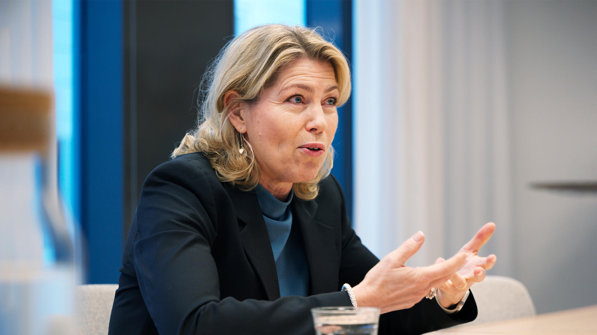 En kvinna i mörkblå blazer presenterar och gestikulerar under ett möte i en modern kontorsmiljö.