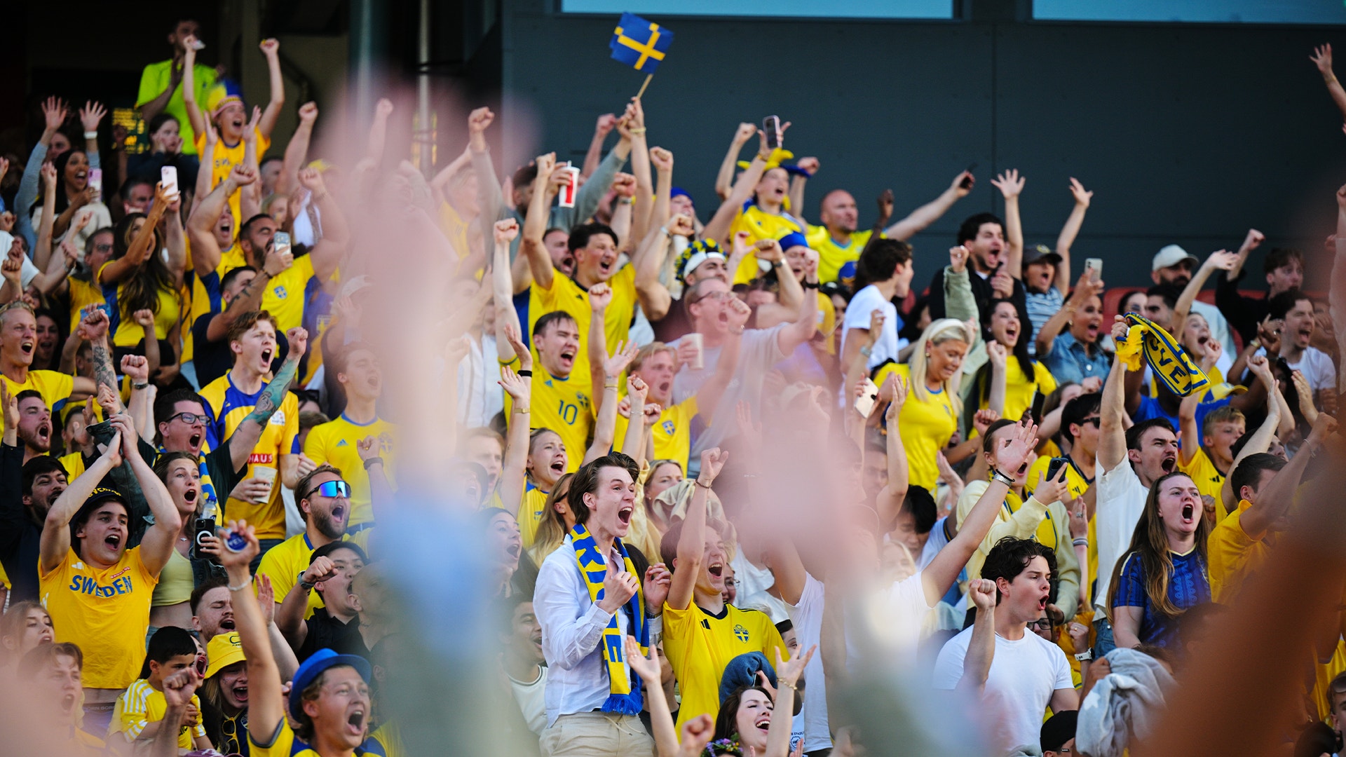En stor grupp fotbollssupportrar i gula och blåa tröjor jublar med armarna upp på ett stadion.