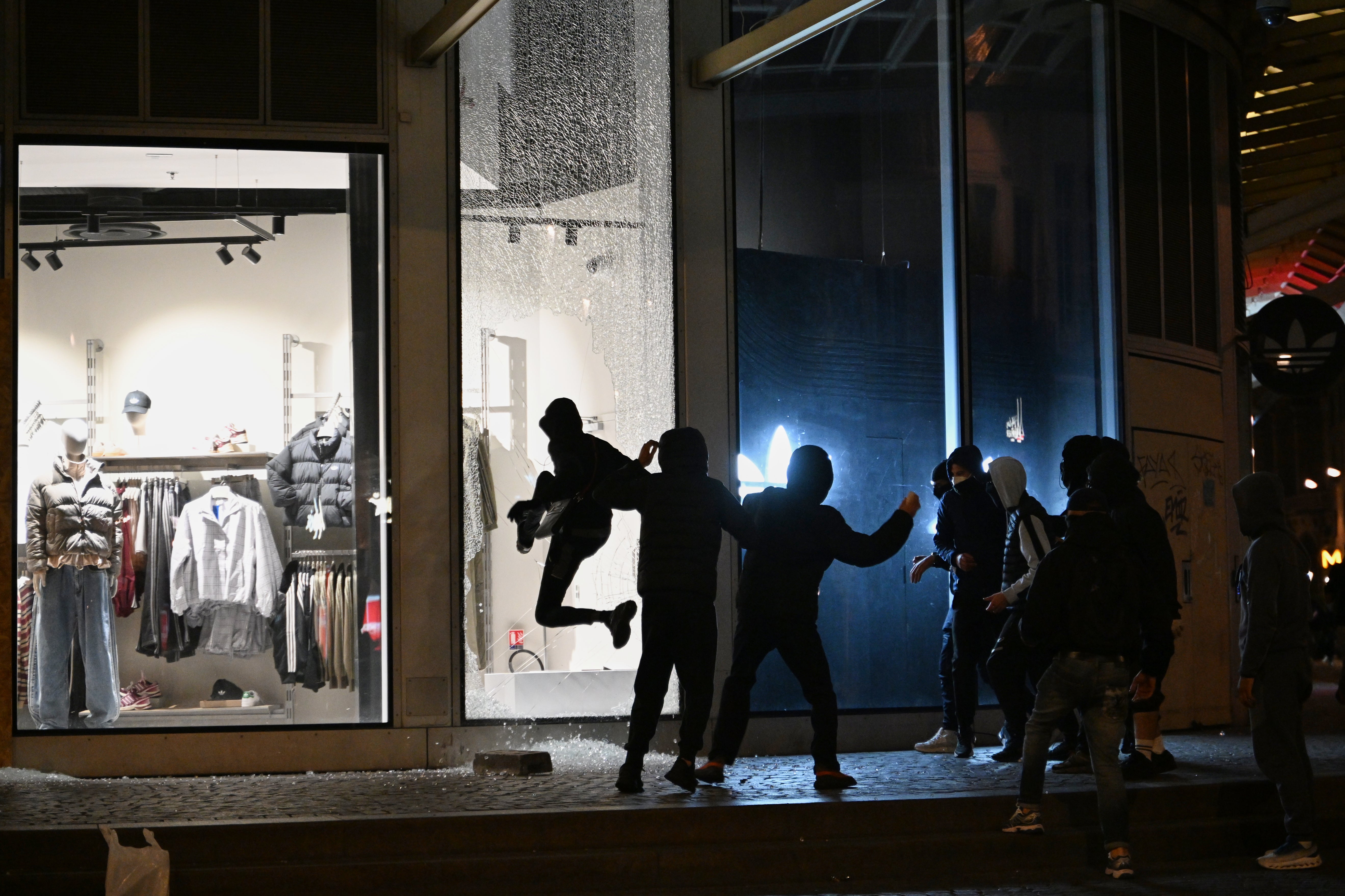 Mörkklädda, maskerade personer slår sönder skyltfönster på en butik i skymningen.