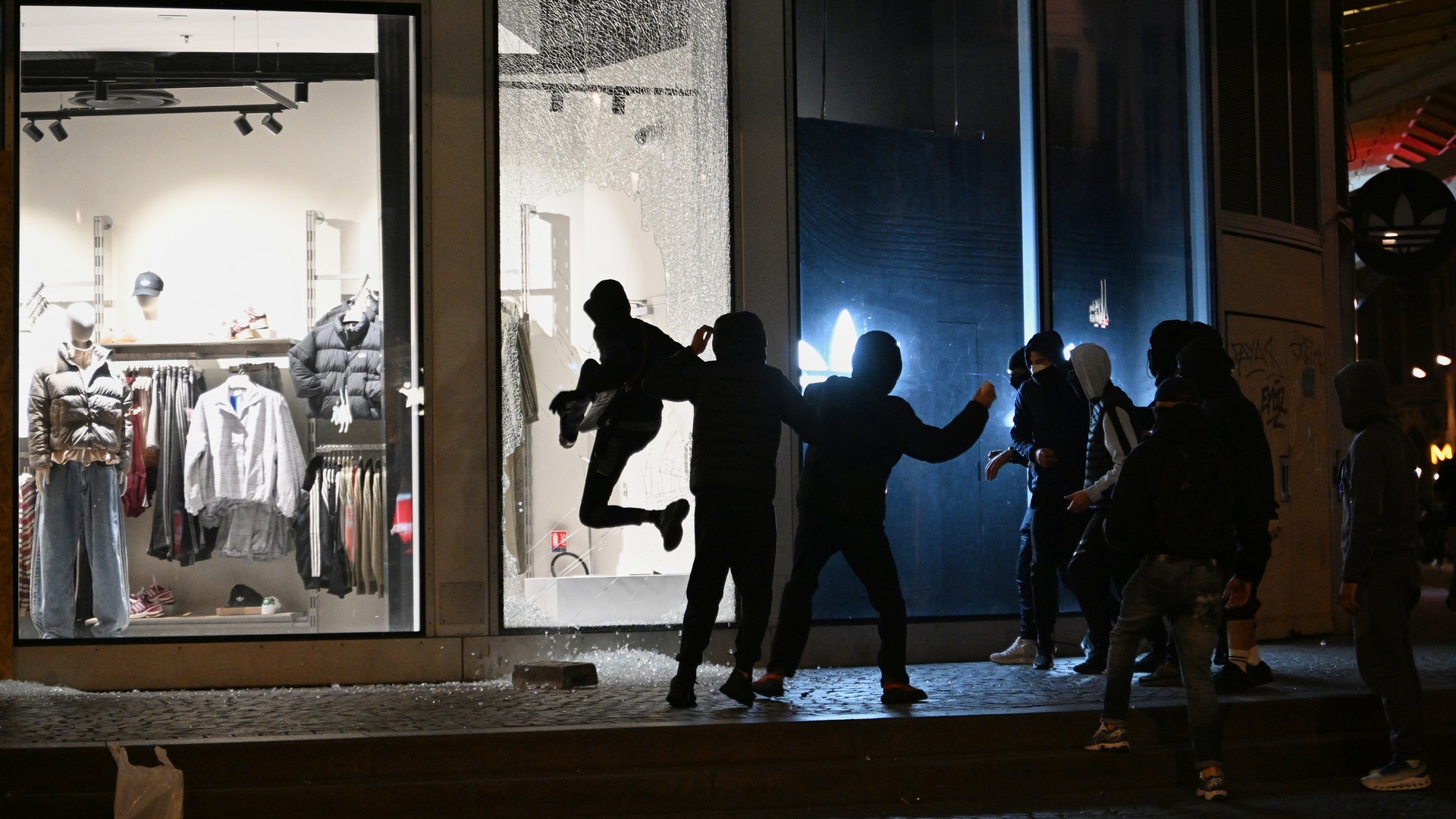 Mörkklädda, maskerade personer slår sönder skyltfönster på en butik i skymningen.