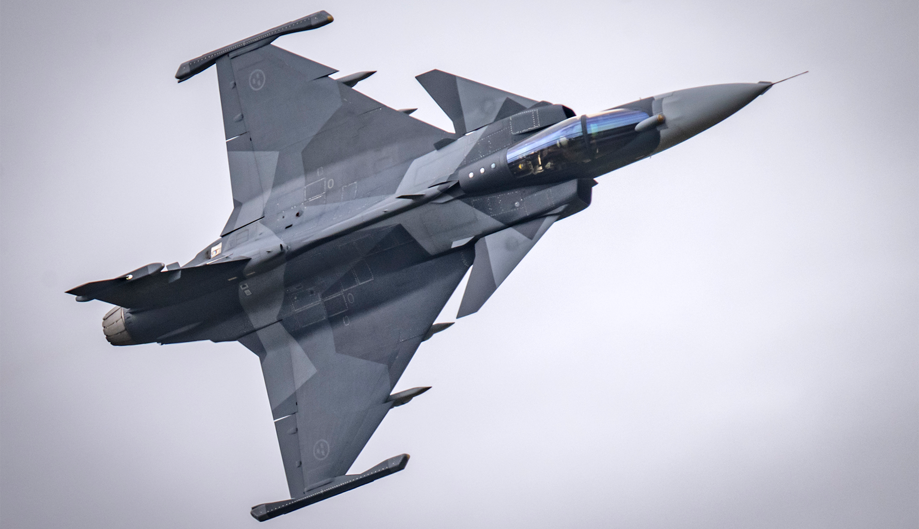 En JAS Gripen stridsflygplan fotograferad från ovan under flygning mot en ljus himmel.