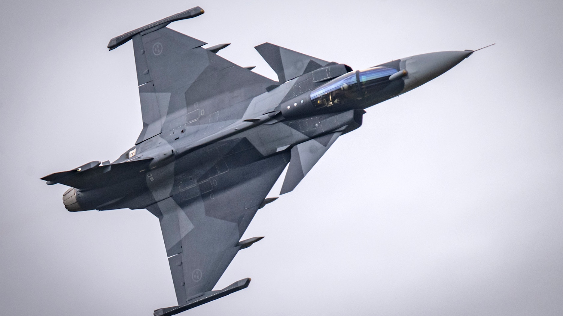 En JAS Gripen stridsflygplan fotograferad från ovan under flygning mot en ljus himmel.