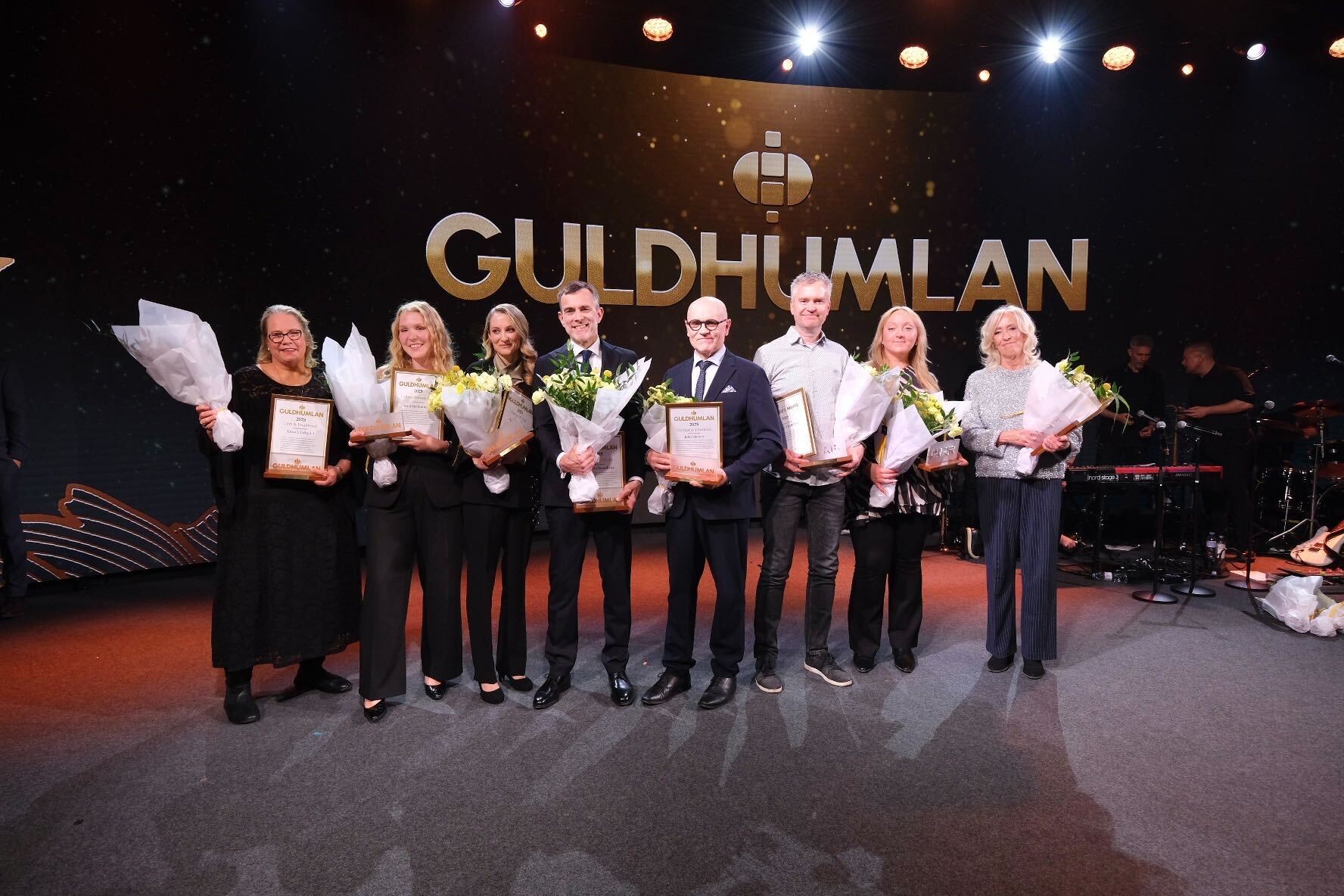 Grupp av människor på en scen vid en ceremoni. De håller blommor och diplom. I bakgrunden syns Guldhumlan-logotypen på en stor skärm.