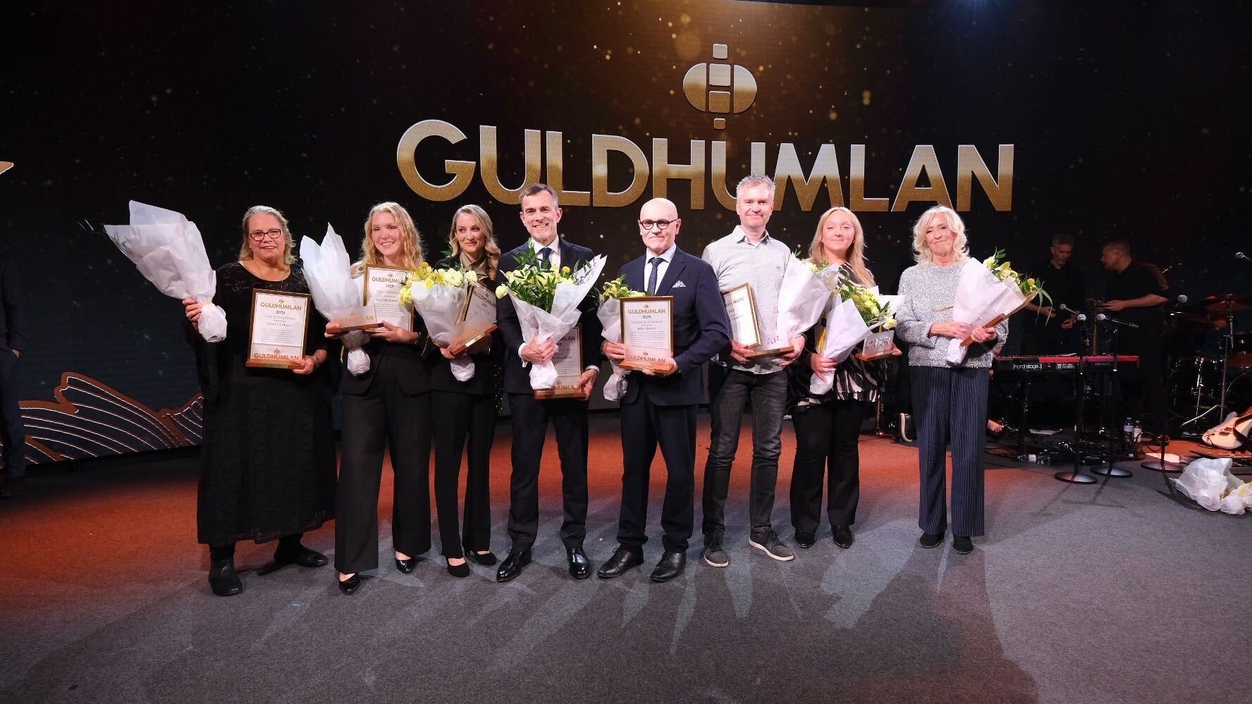 Grupp av människor på en scen vid en ceremoni. De håller blommor och diplom. I bakgrunden syns Guldhumlan-logotypen på en stor skärm.