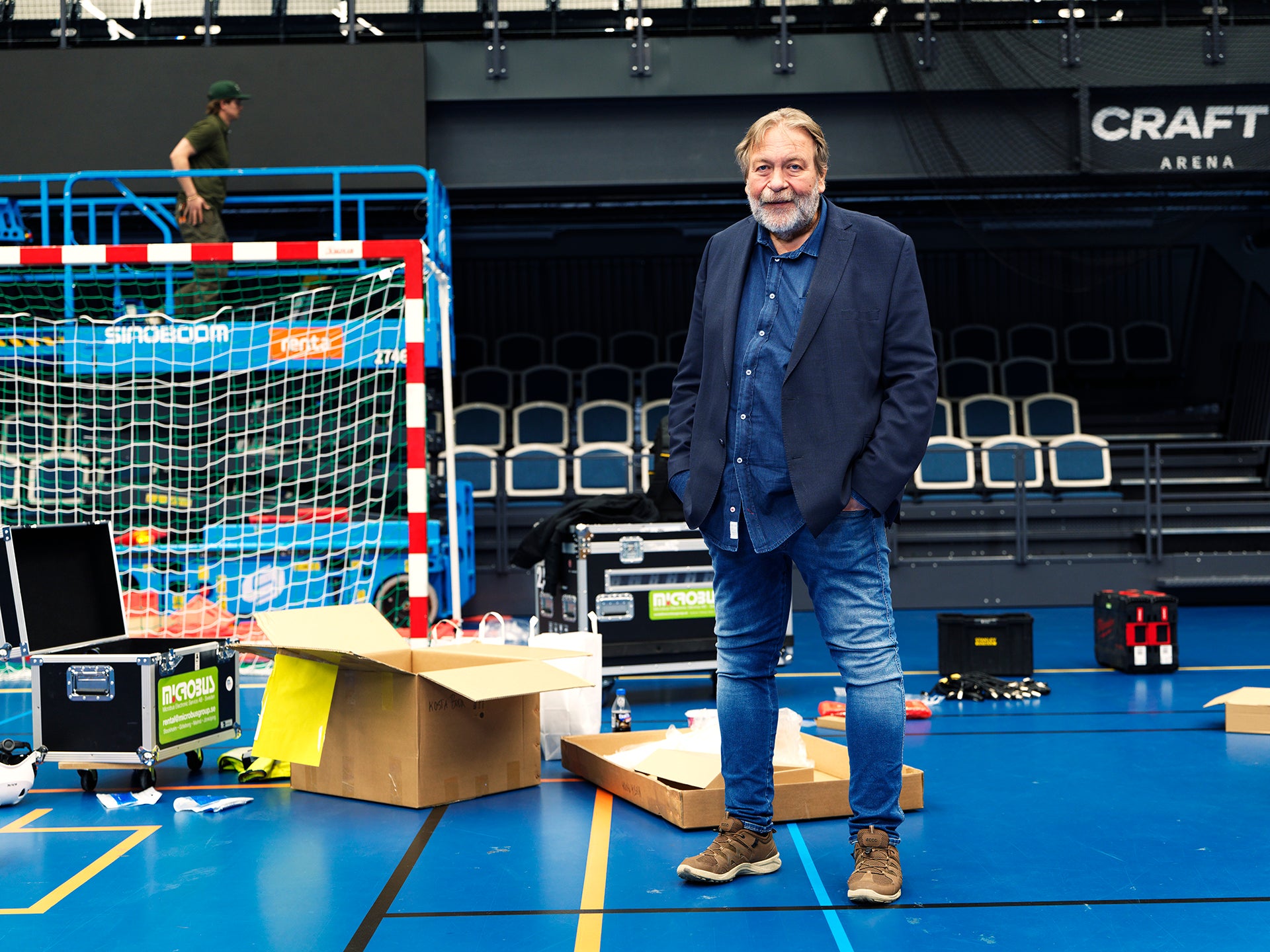 En man i mörkblå jacka står på en blå inomhussporthall med handbolls-/futsal-utrustning omkring sig.