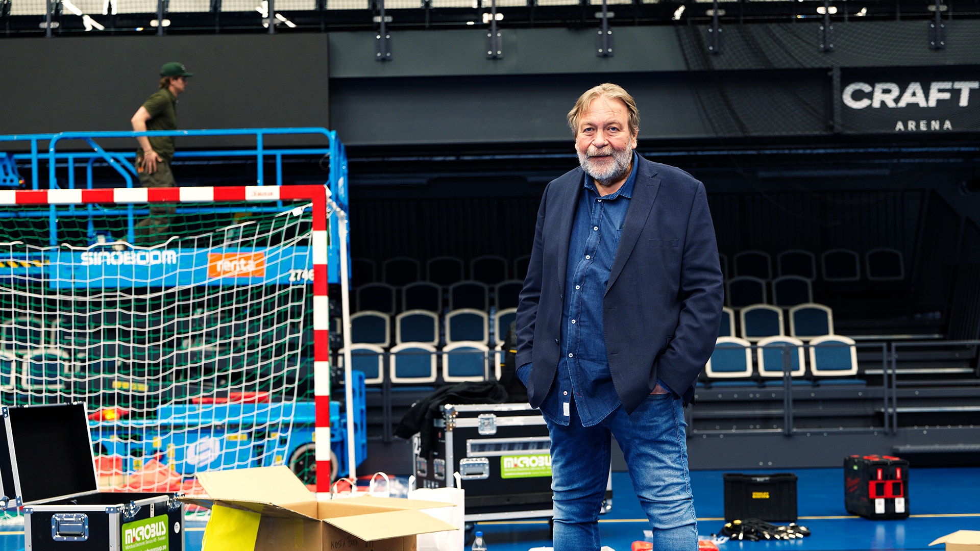 En man i mörkblå jacka står på en blå inomhussporthall med handbolls-/futsal-utrustning omkring sig.