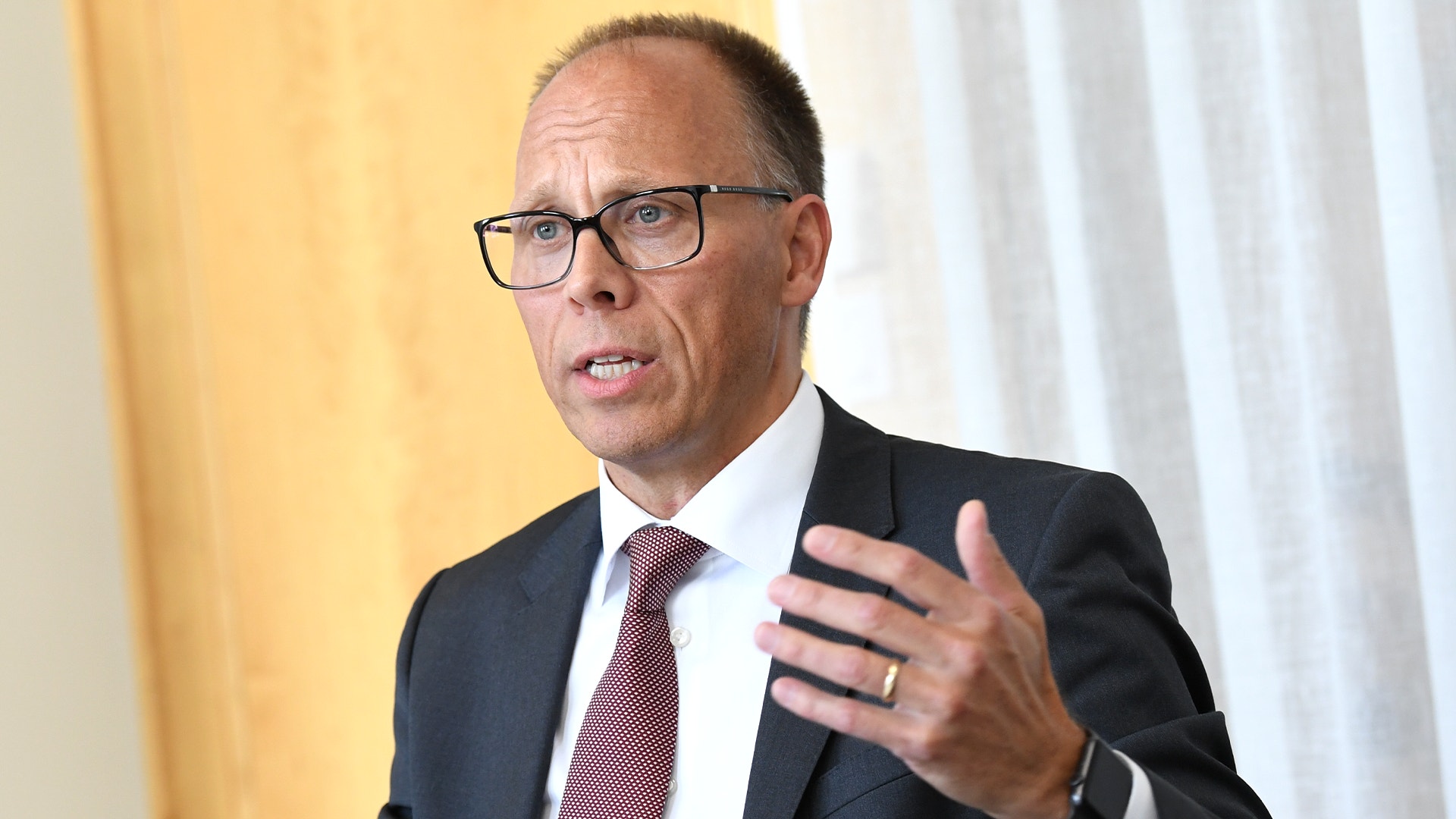 Porträtt av en man i glasögon och kostym som gestikulerar medan han talar.