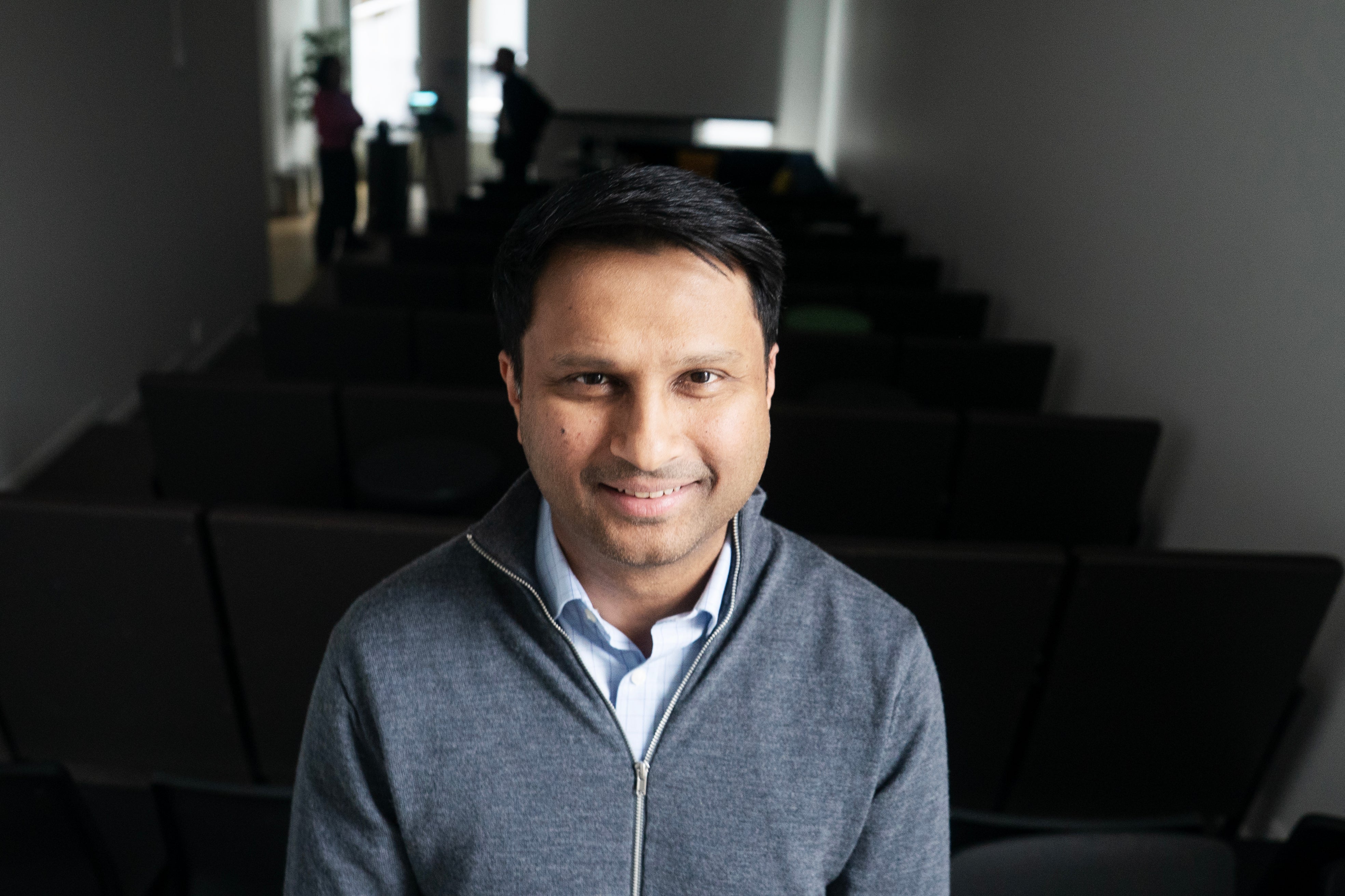 Porträttfoto av Rishit Jhunjhunwala, VD för Truecaller, som ler mot kameran. Han bär en grå cardigan över en vit skjorta och fotograferas inomhus mot en suddig bakgrund med moderna arkitektoniska element.