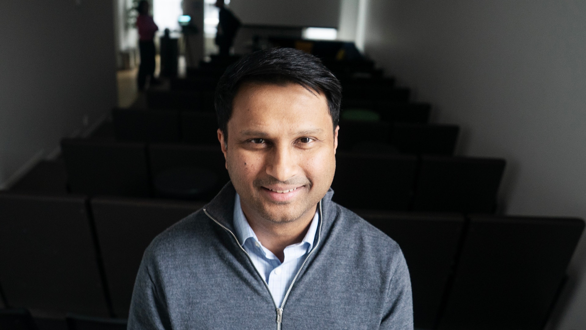 Porträttfoto av Rishit Jhunjhunwala, VD för Truecaller, som ler mot kameran. Han bär en grå cardigan över en vit skjorta och fotograferas inomhus mot en suddig bakgrund med moderna arkitektoniska element.