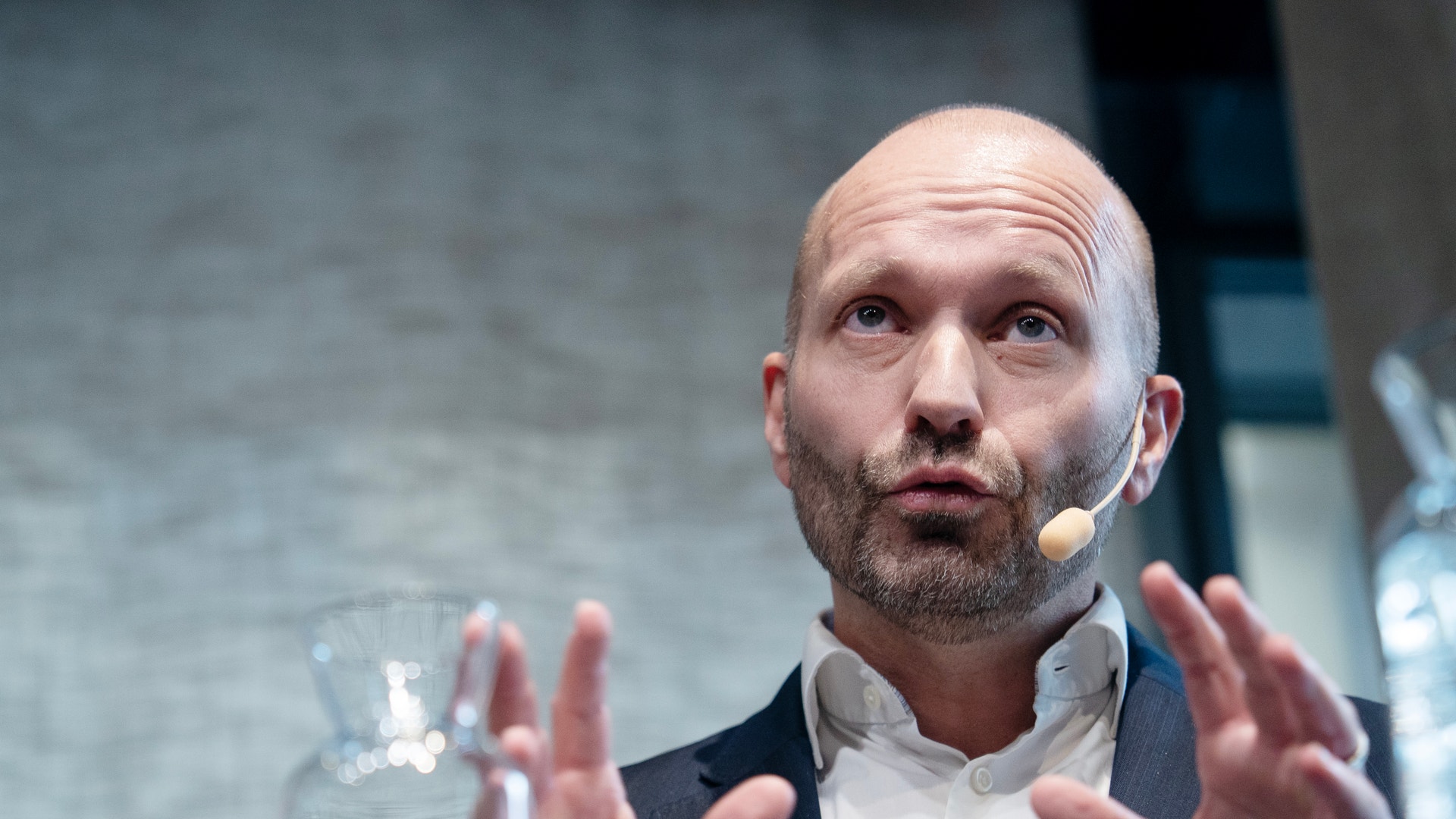 En man med kort skägg och headset talar och gestikulerar med båda händerna under ett seminarium.