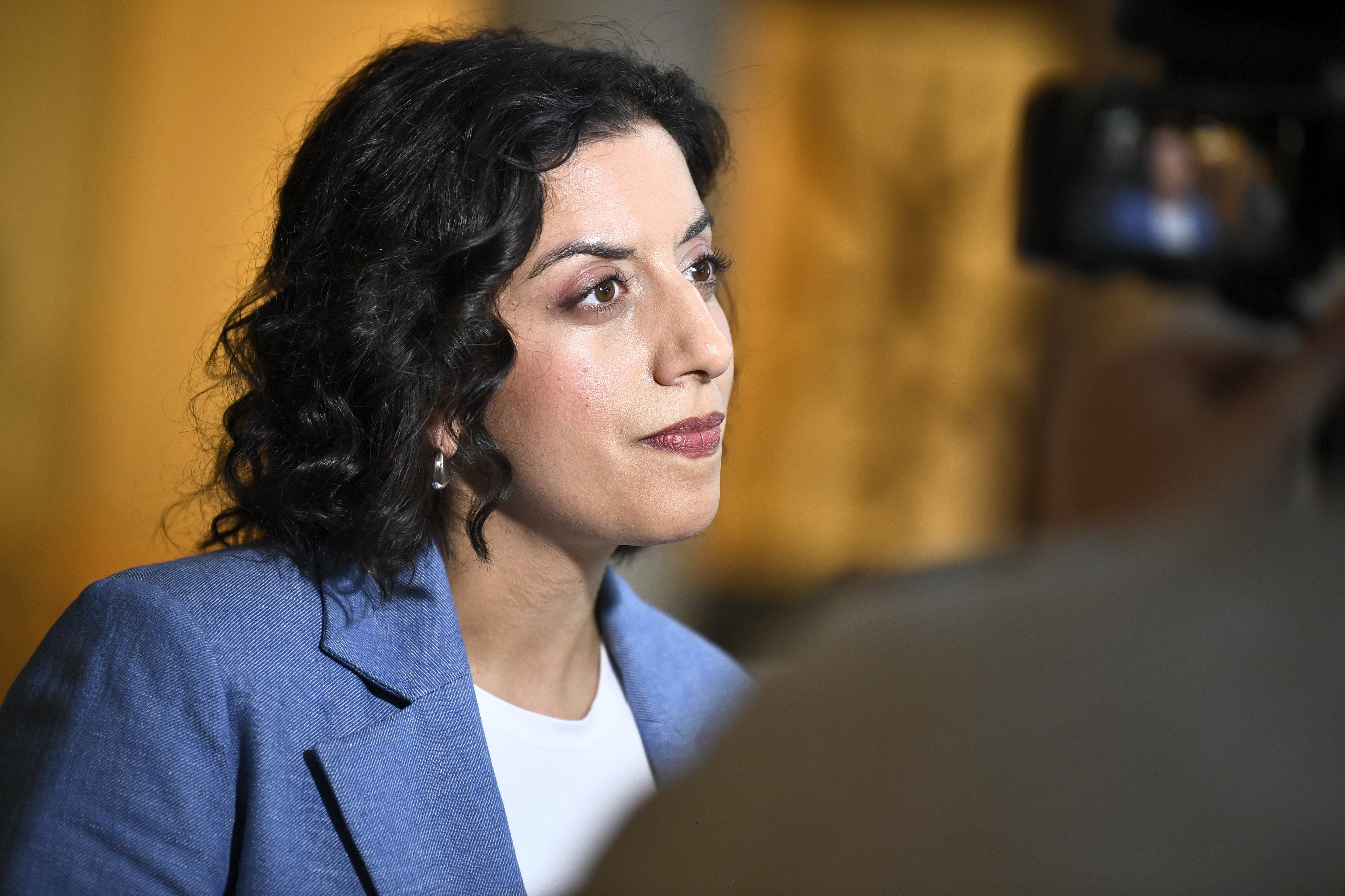 Porträtt av en kvinna med mörkt lockigt hår i blå blazer under ett medieevent eller presssamtal.