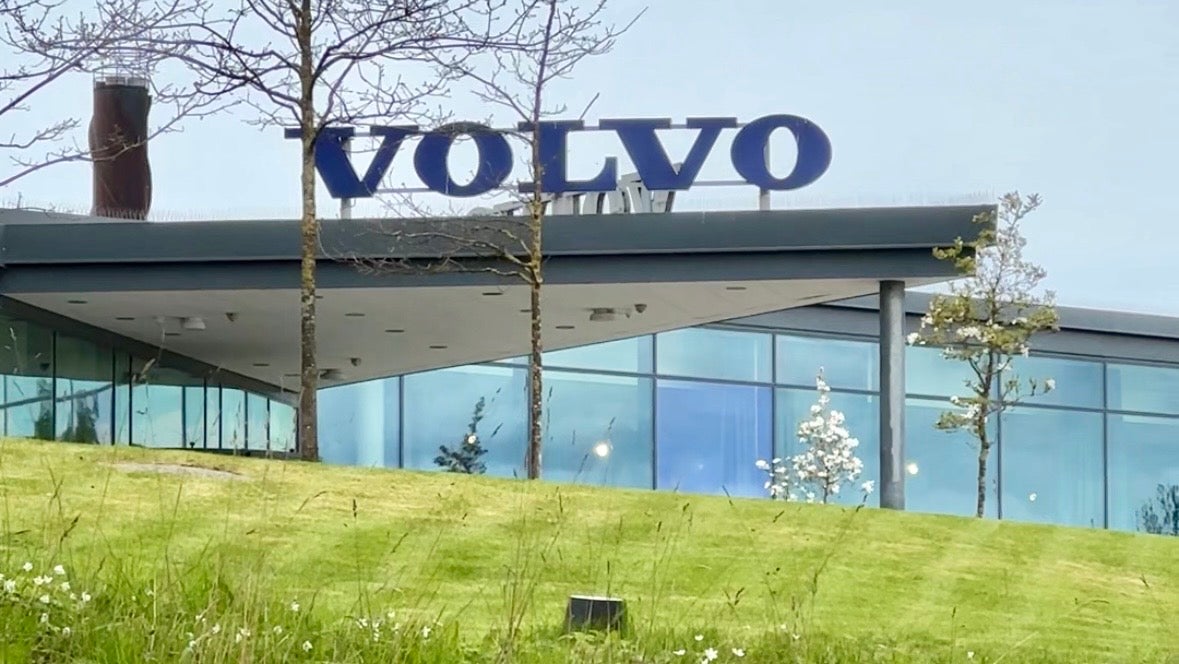 En modern byggnad med Volvo-logotypen på taket. Byggnaden har stora glasfönster och ligger på en grön sluttning med träd.