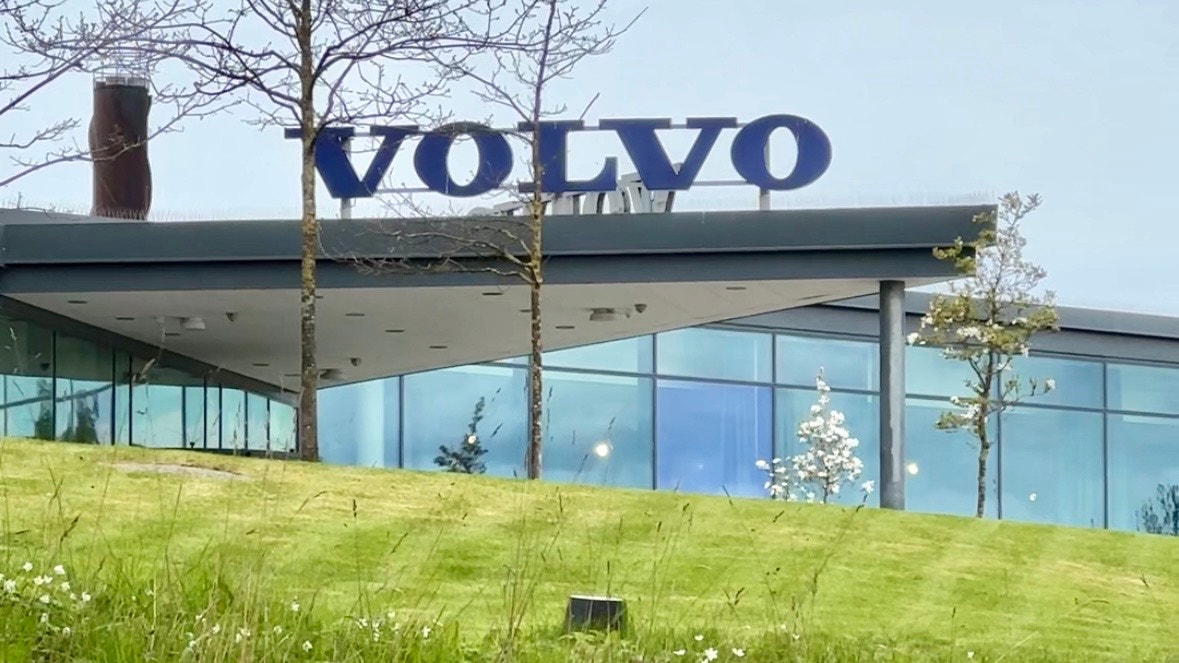 En modern byggnad med Volvo-logotypen på taket. Byggnaden har stora glasfönster och ligger på en grön sluttning med träd.