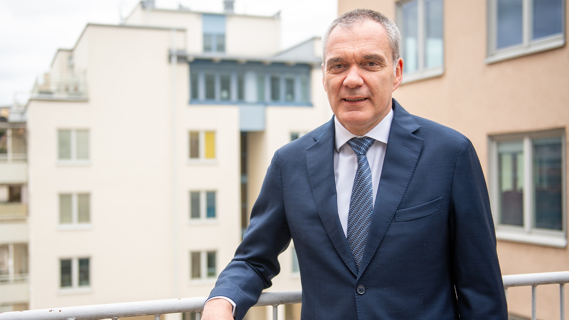 En leende man i mörkblå kostym, vit skjorta och slips står lutad mot ett räcke utomhus. Bakom honom syns bostadshus.