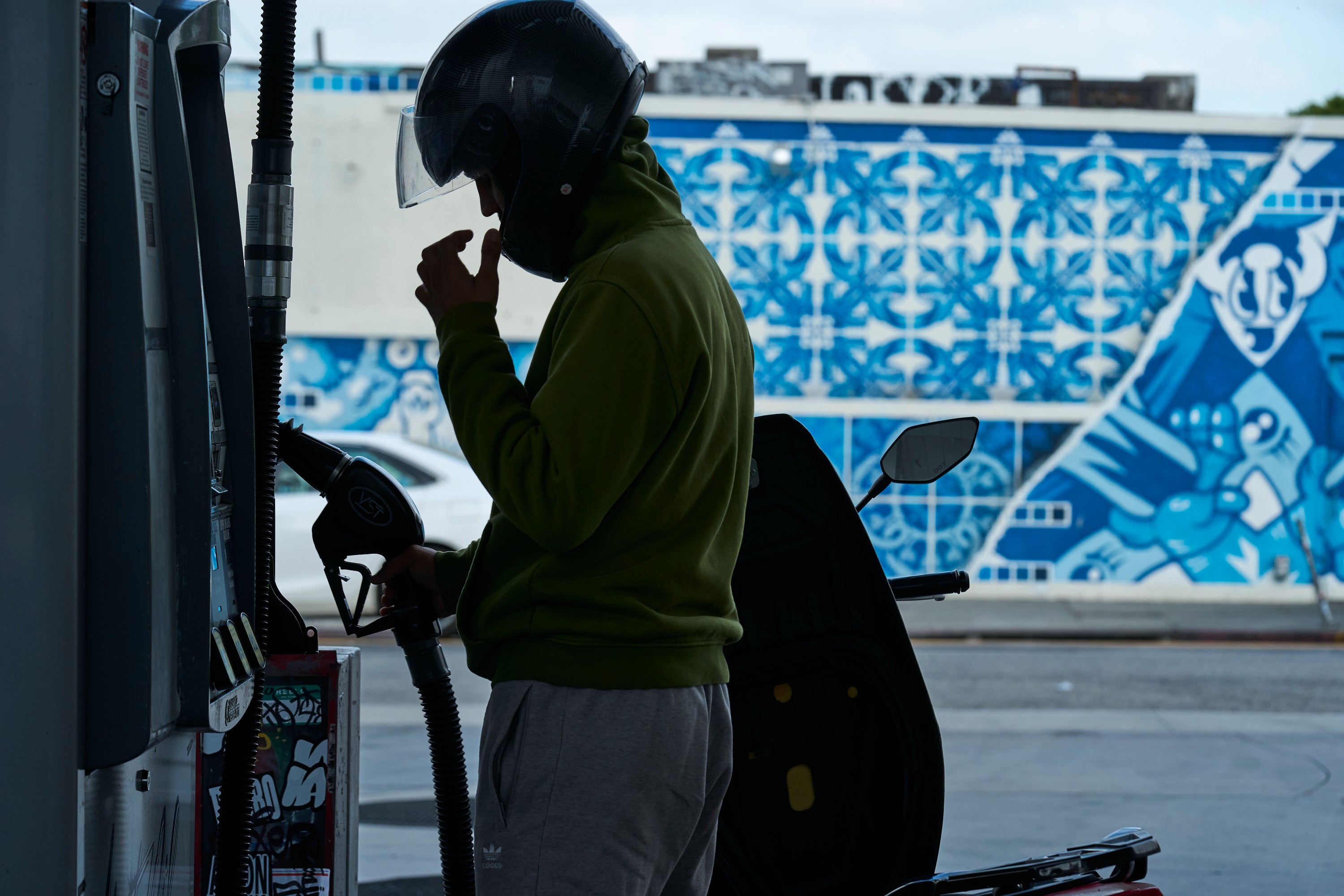 En person i grön jacka och svart hjälm tankar en motorcykel vid en bensinstation. En blå vägg med dekorativt mönster syns i bakgrunden.