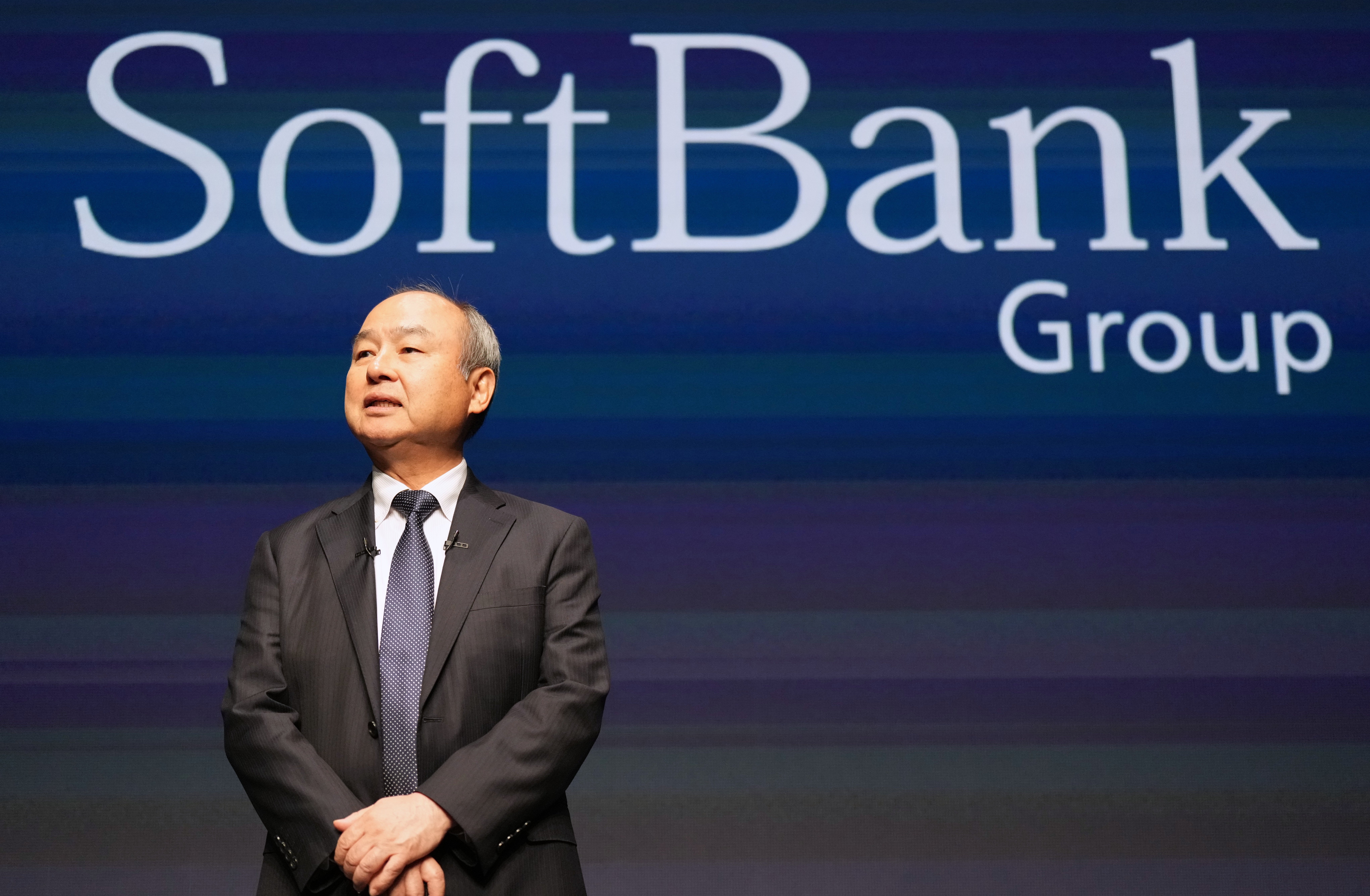 Masayoshi Son i mörk kostym och slips står framför en skärm med texten SoftBank Group.