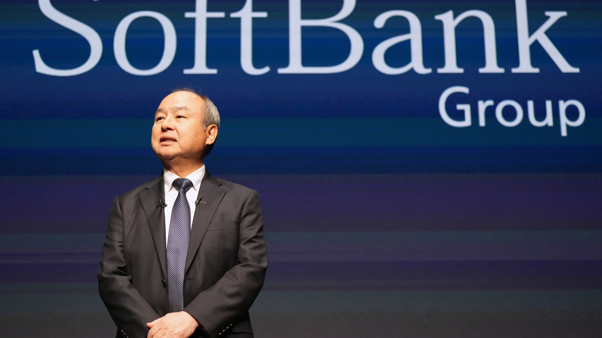 Masayoshi Son i mörk kostym och slips står framför en skärm med texten SoftBank Group.