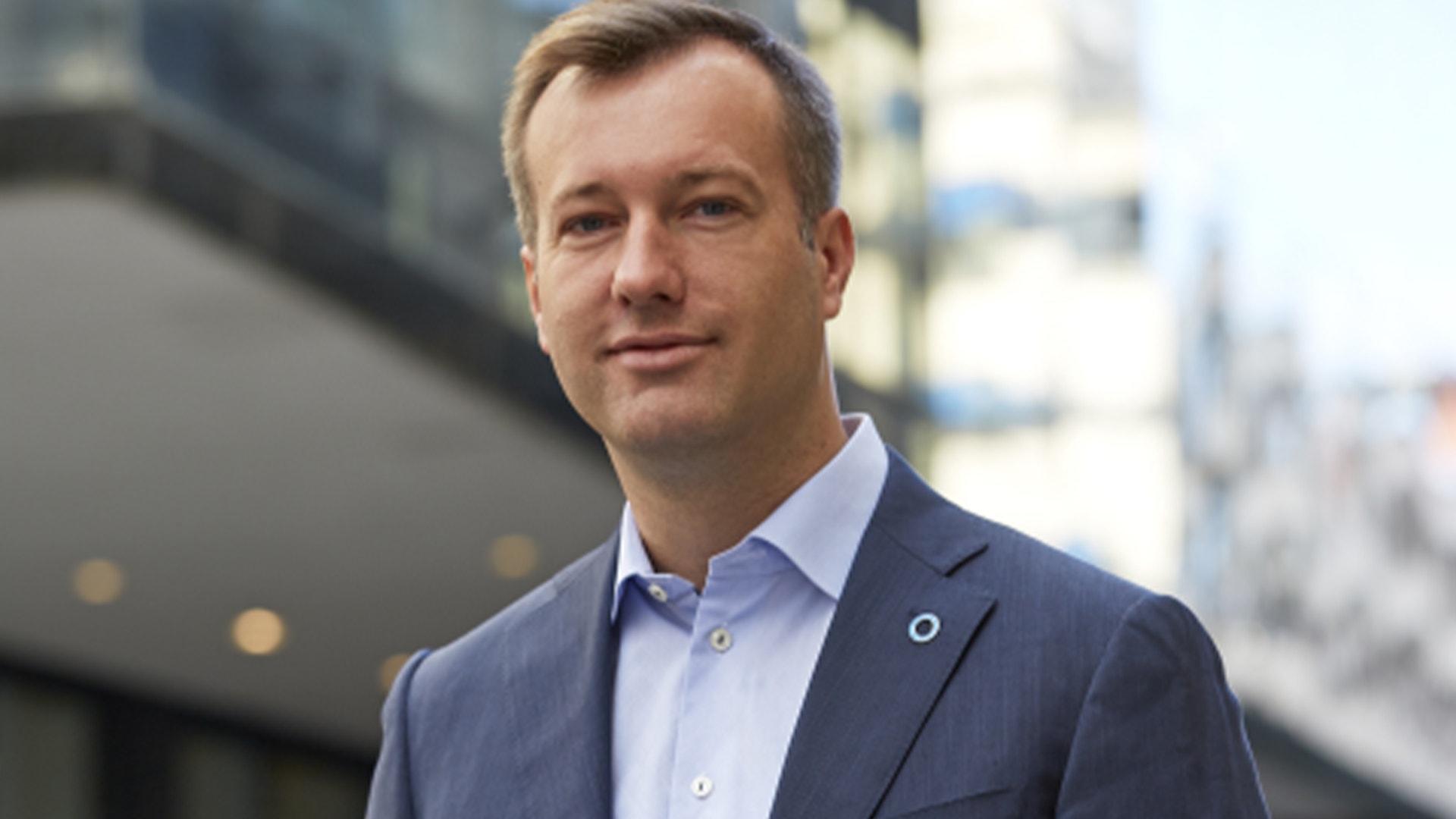 Porträtt av en man i blå kavaj och ljusblå skjorta framför en urban bakgrund.