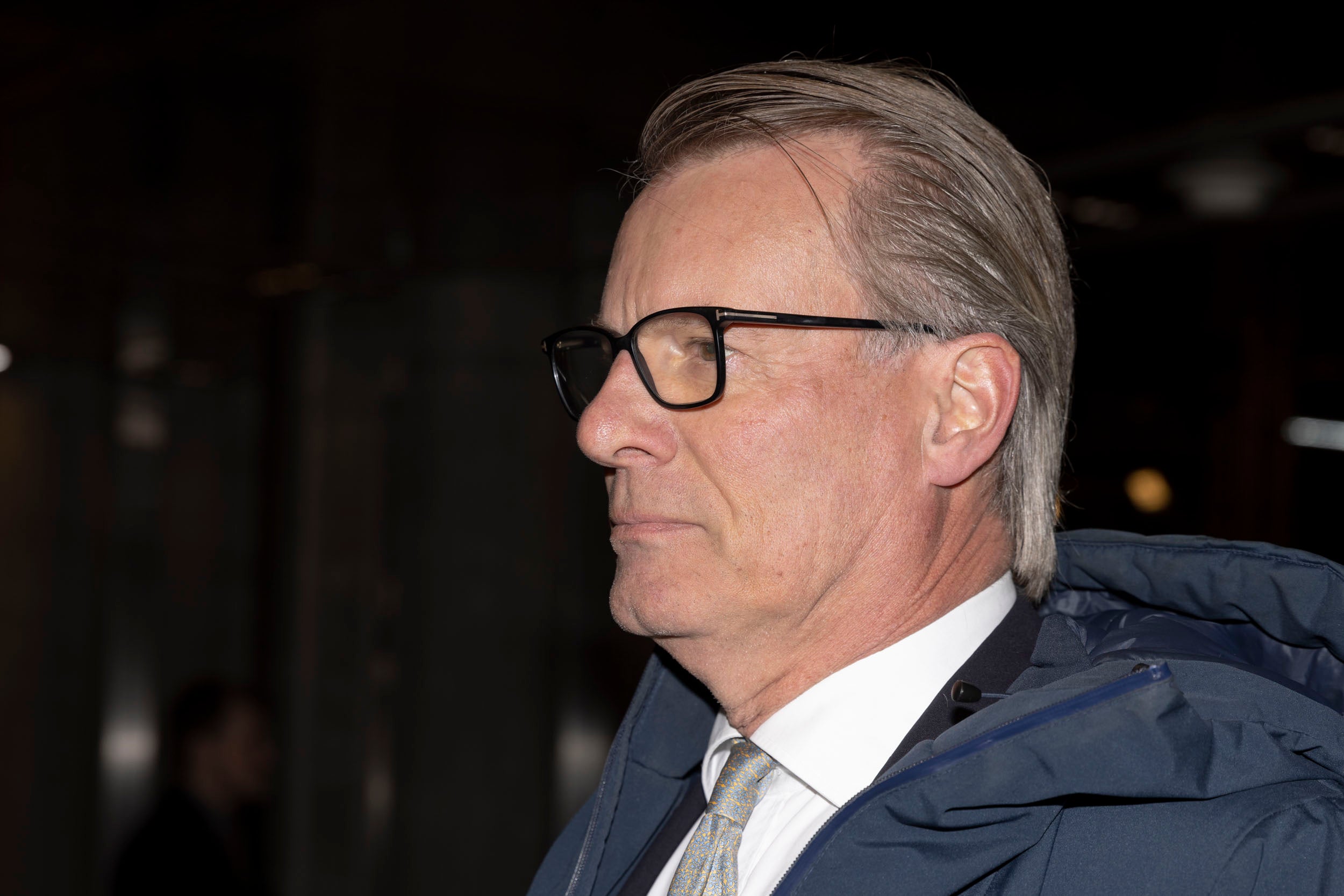 Porträtt i profil av en man med glasögon och bakåtkammat grått hår. Han bär en mörkblå jacka, vit skjorta och grå slips.