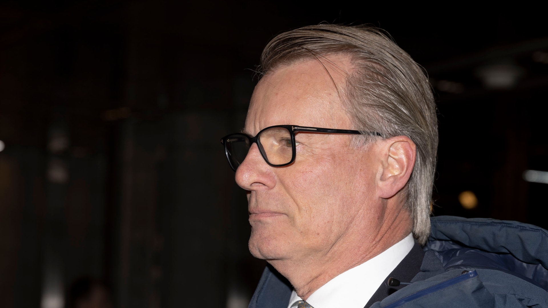 Porträtt i profil av en man med glasögon och bakåtkammat grått hår. Han bär en mörkblå jacka, vit skjorta och grå slips.