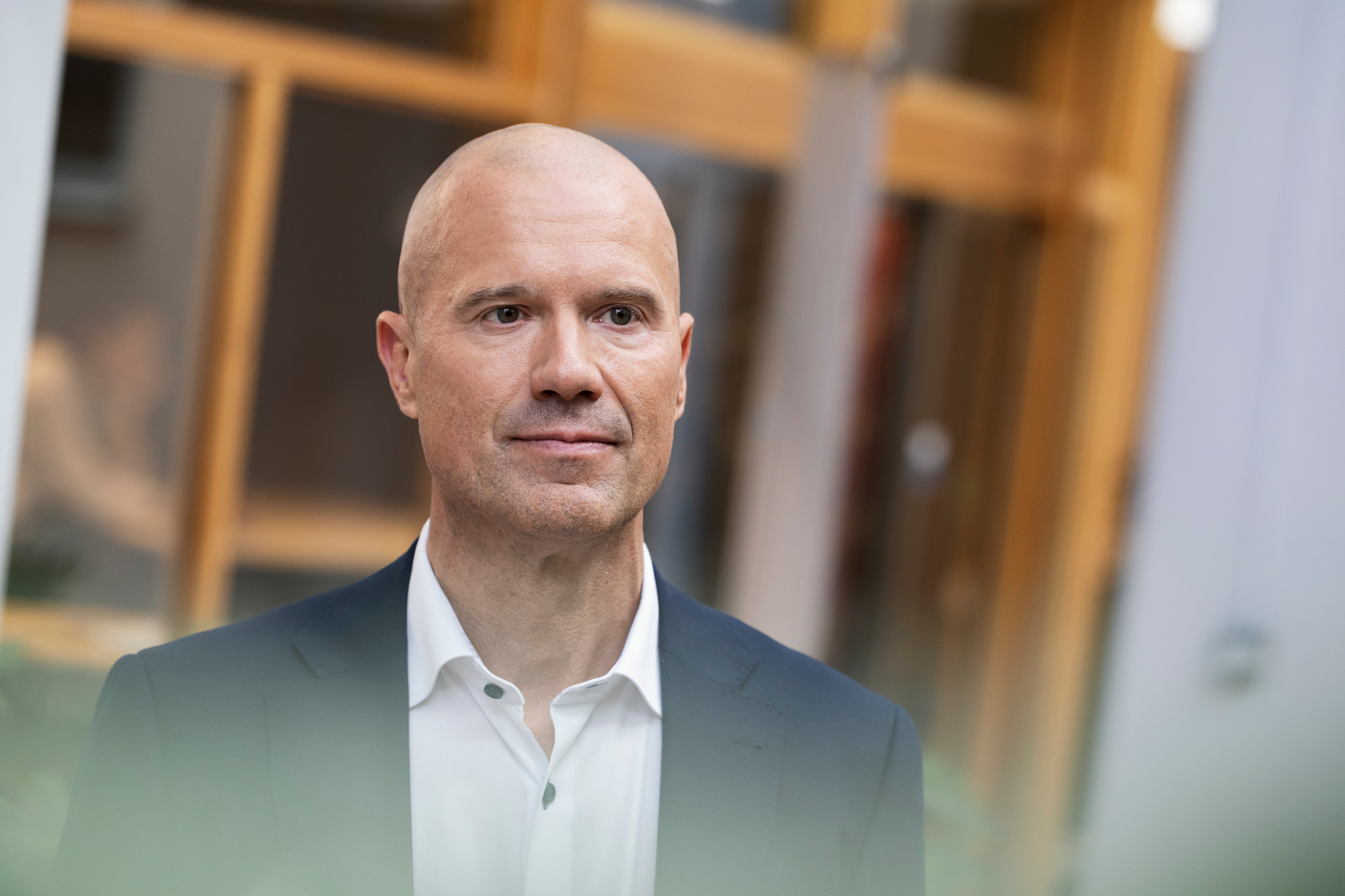 Porträtt av en man i mörk blazer och vit skjorta framför en suddig bakgrund med träfärgade dörrar.