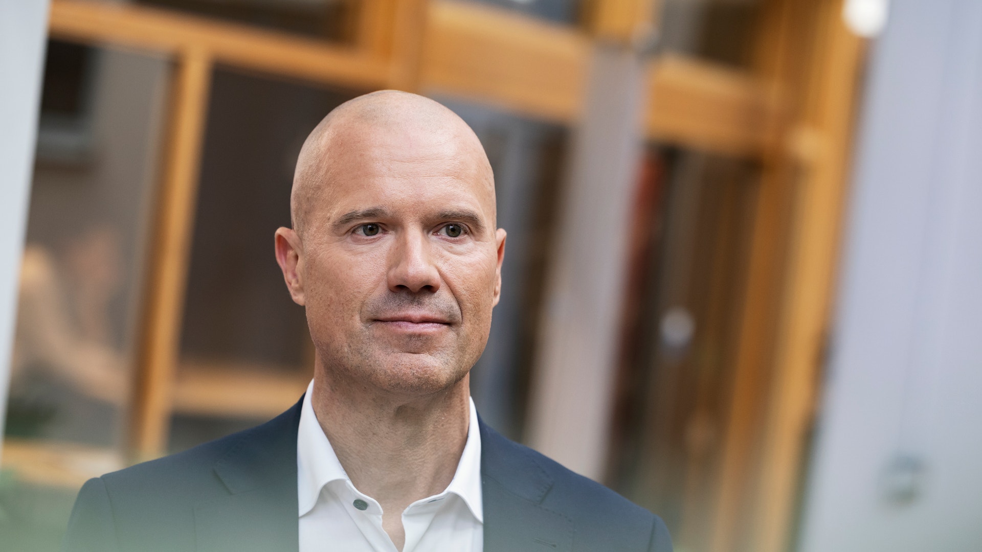 Porträtt av en man i mörk blazer och vit skjorta framför en suddig bakgrund med träfärgade dörrar.
